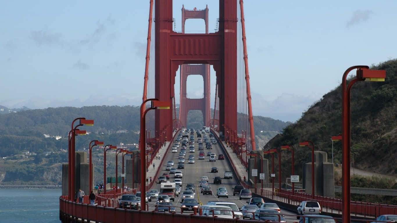 1366x768 San Francisco 桌布 壁紙 — 一座繁忙的紅色吊橋橫跨海灣，汽車在晴朗的藍天和綠色的山坡下行駛。