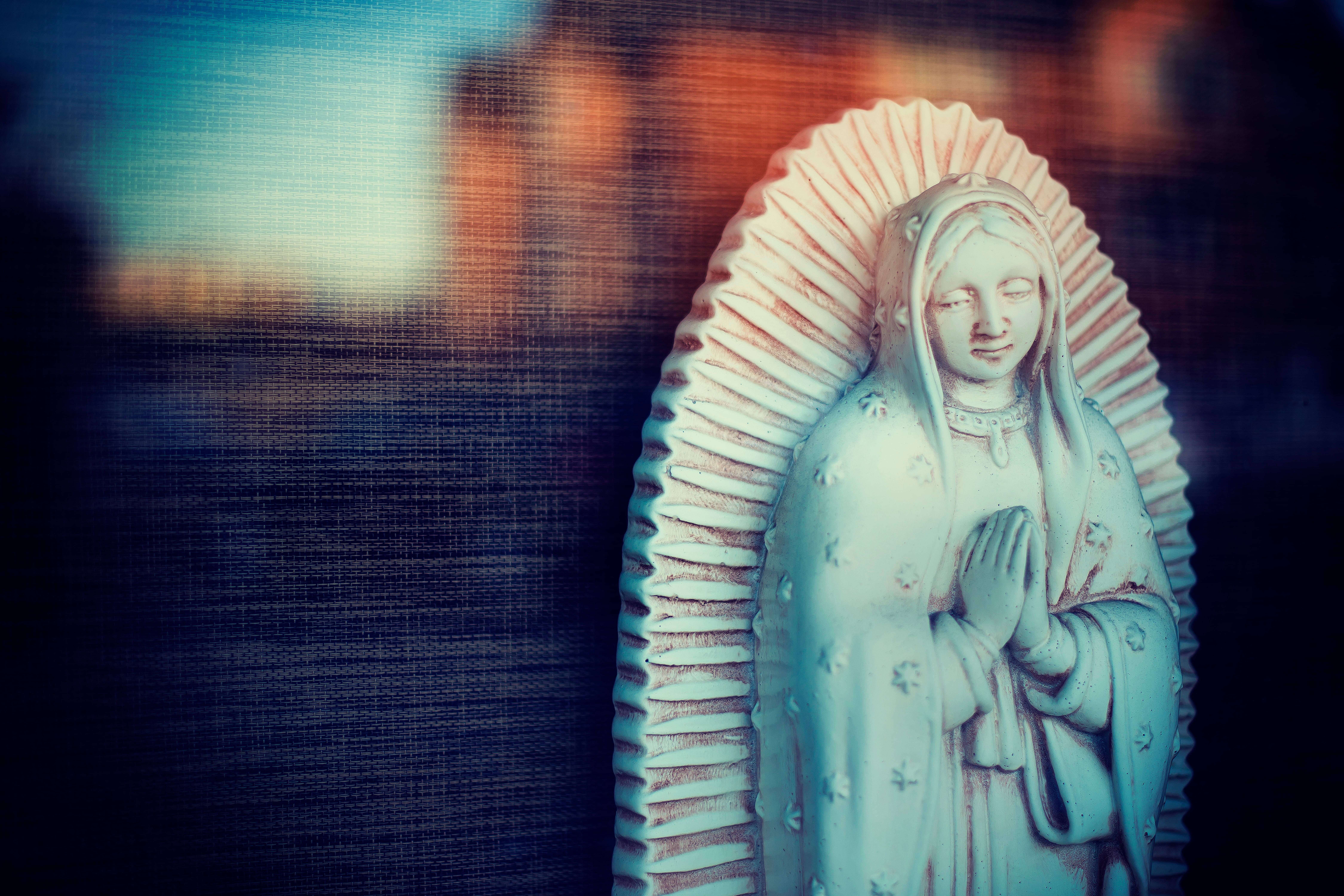 Catholic 桌布 壁紙 — 一座聖母瓜達盧佩的寧靜雕像，雙手合十祈禱，在模糊的暮色城市景觀中柔和發光。