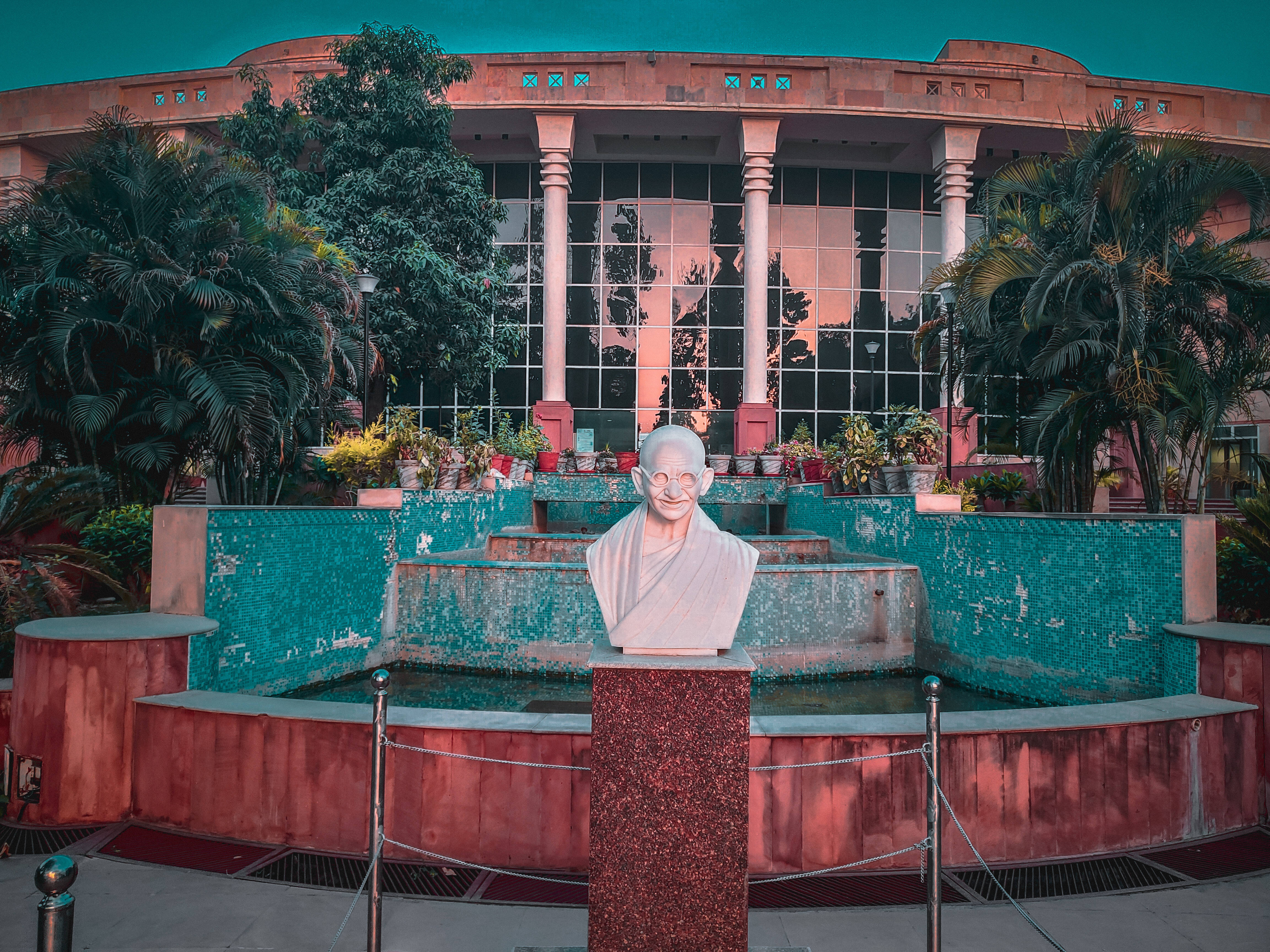 Mahatma Gandhi 桌布 壁紙 — 一座聖雄甘地的大理石半身像矗立在基座上，前方是青綠色瓷磚噴泉，周圍環繞著茂密的棕櫚樹和一座帶玻璃窗的現代建築。