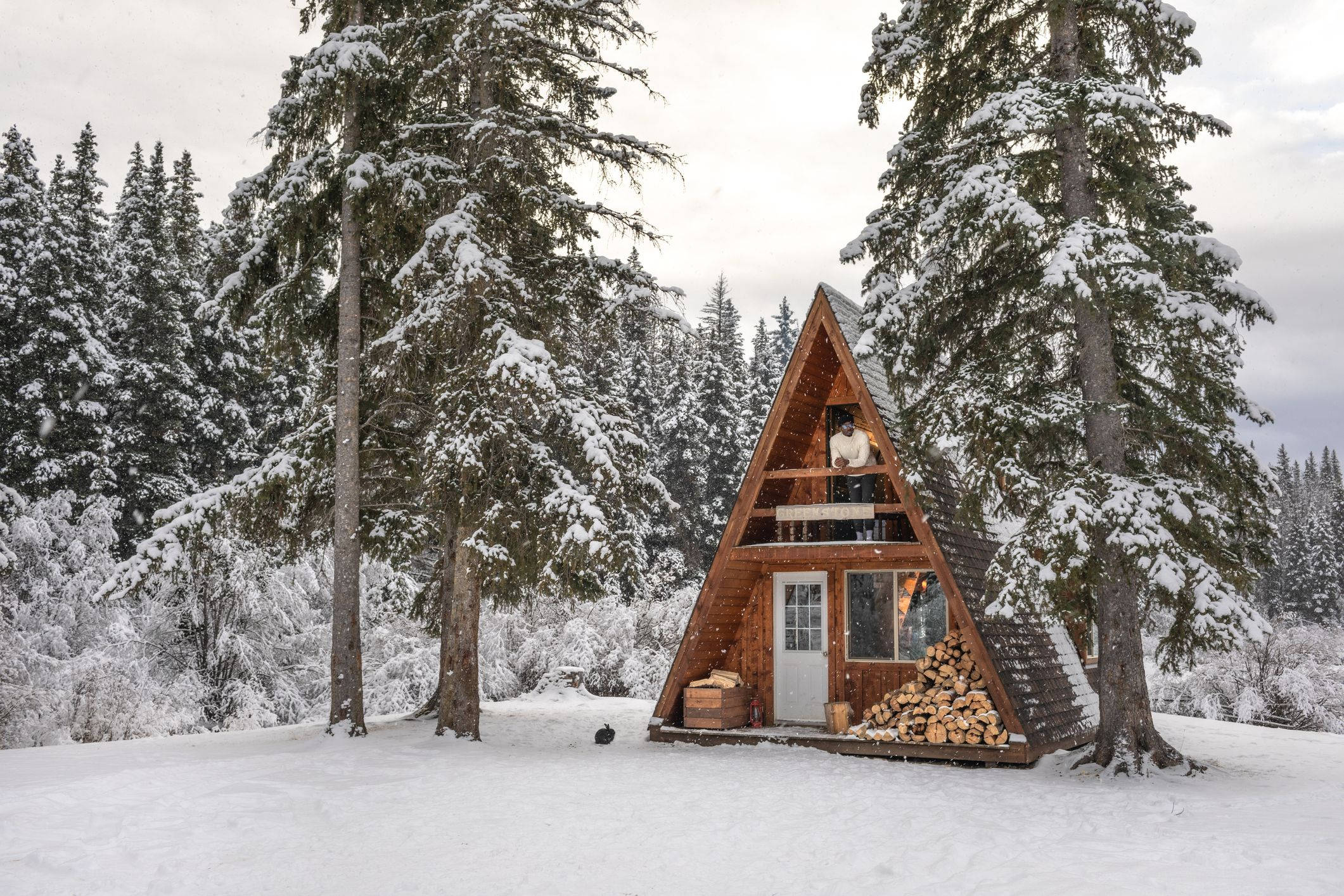 Winter House 桌布 壁紙 — 一座舒適的木製A字型小屋，坐落在雪地森林中，周圍是白雪覆蓋的松樹，陽台上有一個人。
