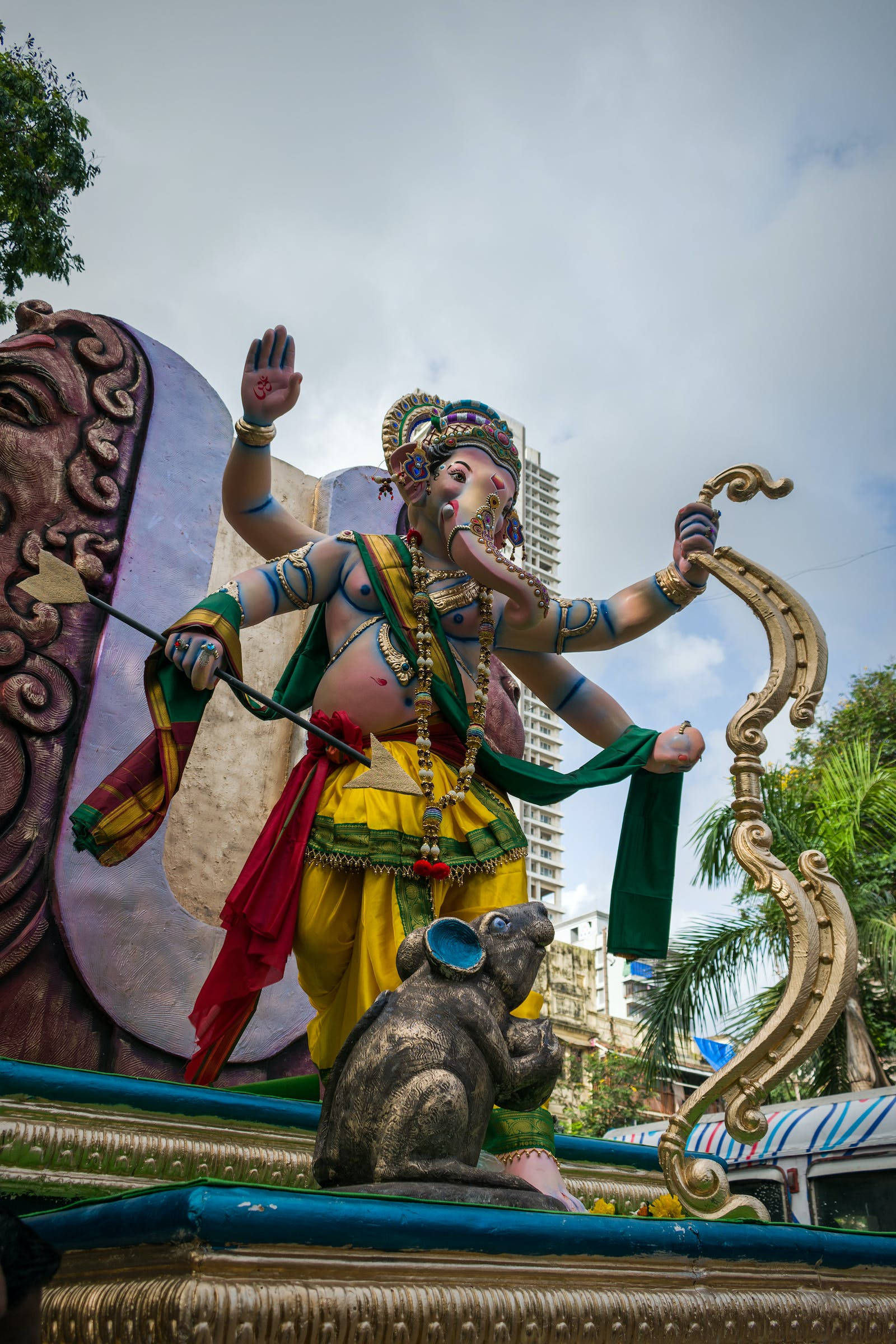 Ganesh 桌布 壁紙 — 一座色彩繽紛、多臂的象神雕像穿著鮮豔服飾，站在裝飾華麗的花車上，旁邊有一隻老鼠同伴，背景是陰天和城市建築。