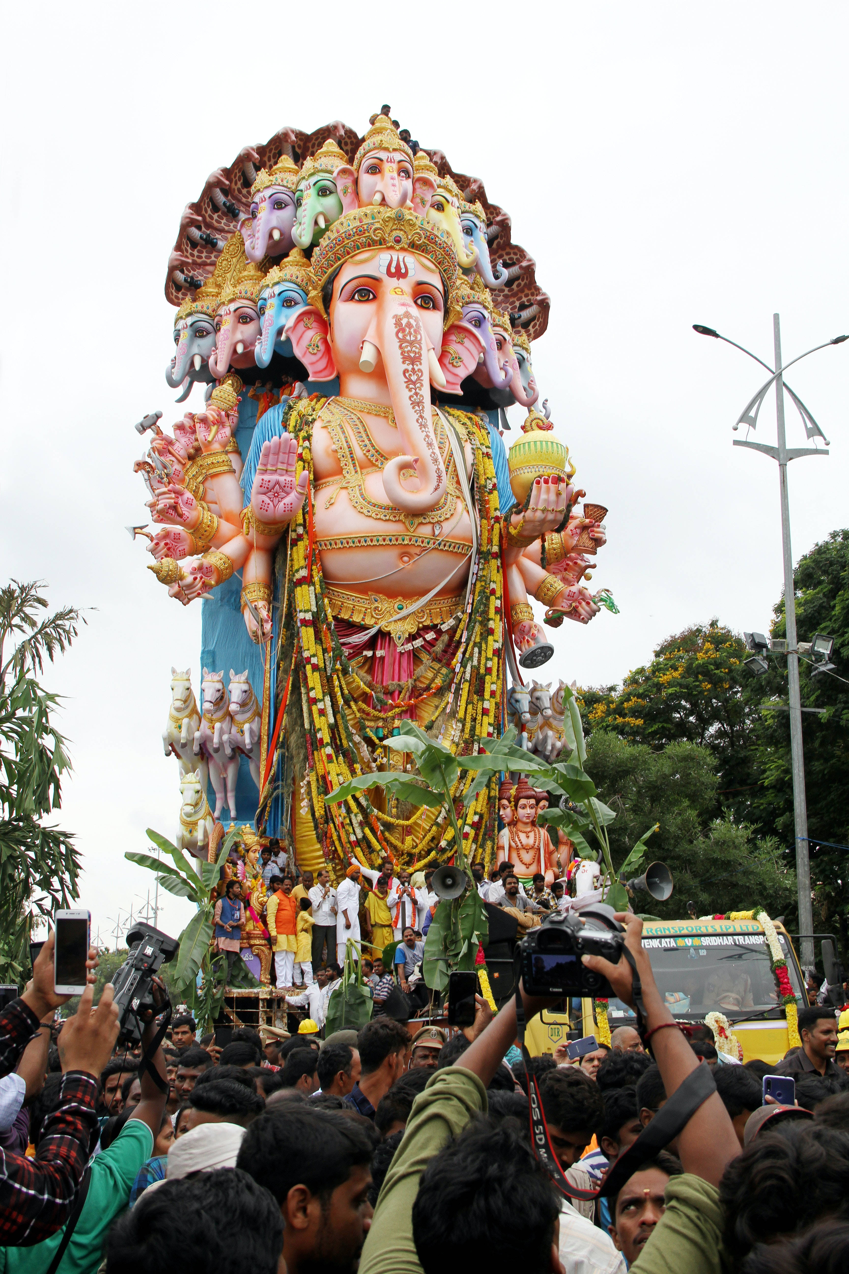 Ganesh Chaturthi 桌布 壁紙 — 一座色彩繽紛、多頭多臂的象神像在擠滿人的街道上遊行，信徒和攝影師在下方聚集。