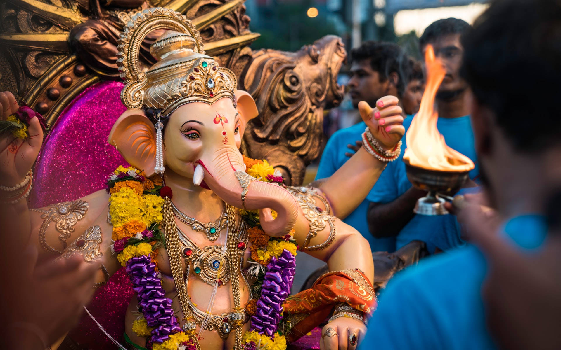 Ganesh 桌布 壁紙 — 一座色彩繽紛、飾有鮮花和珠寶的金色象神雕像，信徒手持點燃的油燈進行禮拜。