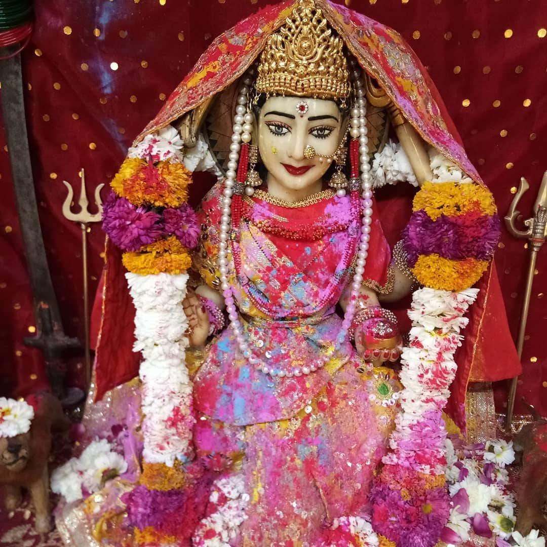 Goddess 桌布 壁紙 — 一座色彩繽紛的印度教女神雕像，披著粉紅色和紅色，戴著金色皇冠，周圍環繞著彩色花環，背景是紅色圓點。