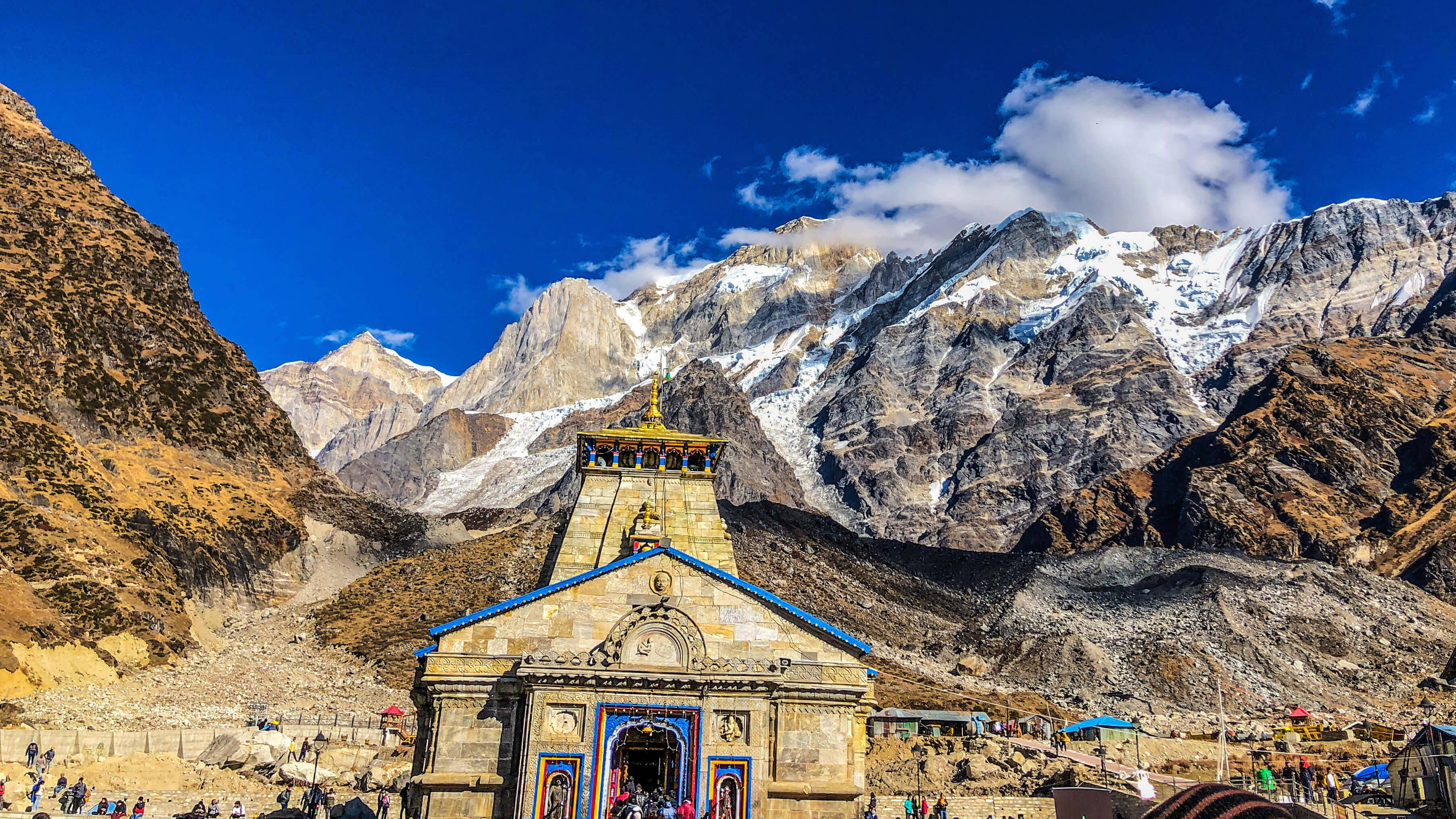 Kedarnath 桌布 壁紙 — 一座色彩繽紛的寺廟，藍色屋頂和鮮豔細節，矗立在晴朗藍天下高聳的雪山前。