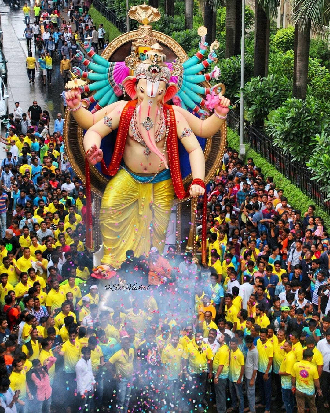 hindu iPhone 桌布 壁紙 — 一座色彩繽紛的巨大象神雕像漂浮在密集的人群上方，人們穿著黃色襯衫，參加充滿活力的節日遊行。