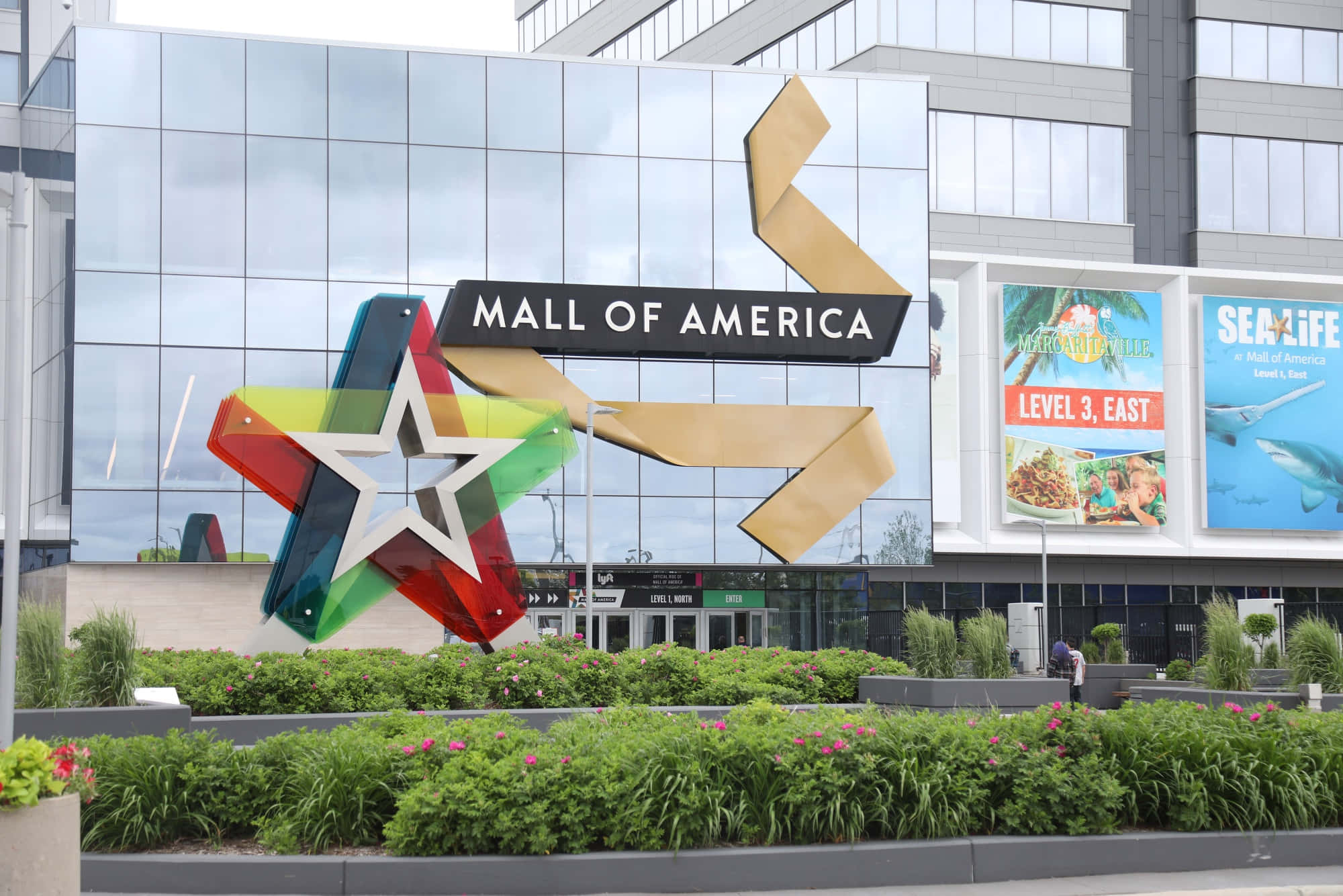 Mall Of America 桌布 壁紙 — 一座色彩繽紛的彩虹星星雕塑矗立在美國購物中心的玻璃幕牆前，周圍環繞著綠植和花壇。