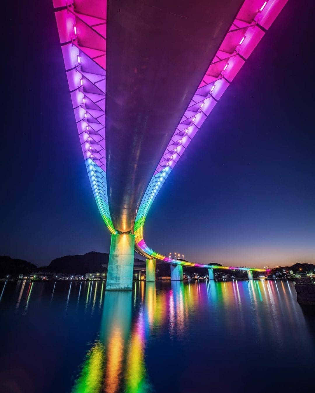 Rainbow Bridge 桌布 壁紙 — 一座色彩繽紛的彩虹燈光橋樑在夜晚橫跨平靜的水面，水面上閃爍著多彩的倒影。