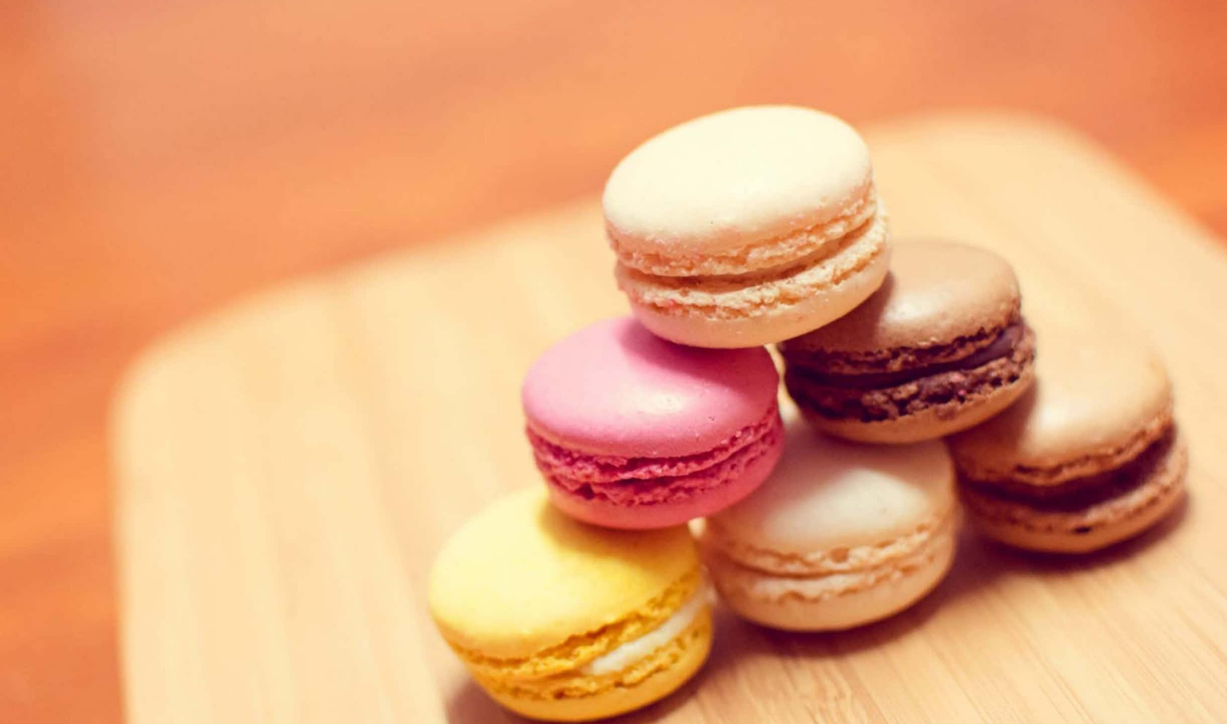 2440x1440 Desserts 桌布 壁紙 — 一座色彩繽紛的馬卡龍金字塔，以柔和粉彩色調呈現，放置在溫暖的木質表面上，背景帶有柔和的散景效果。