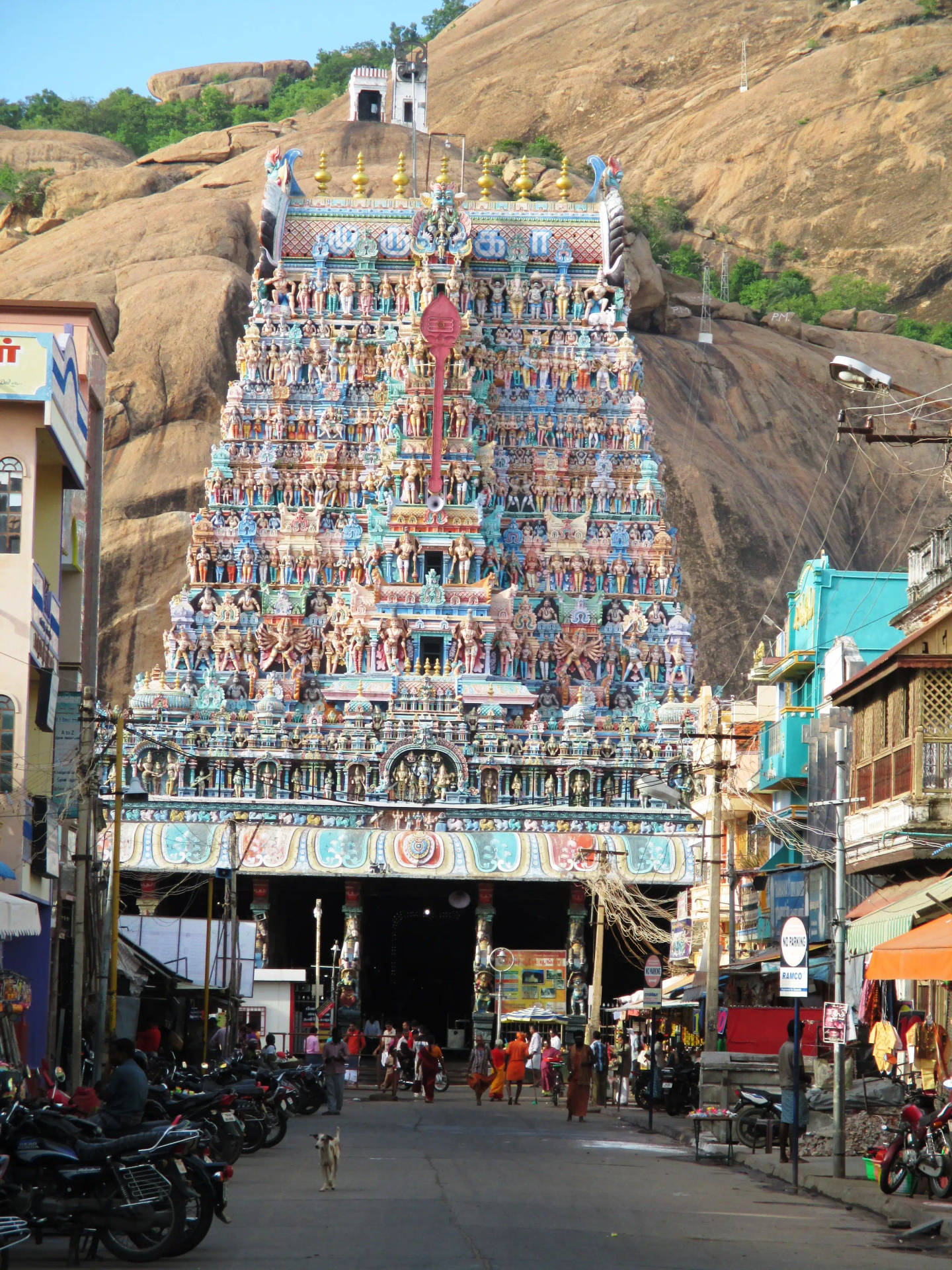 Hindu Temple 桌布 壁紙 — 一座色彩鮮豔、雕刻精美的印度教寺廟高塔（gopuram）雄偉地聳立在岩石山坡上，其底部擠滿了人群和摩托車。