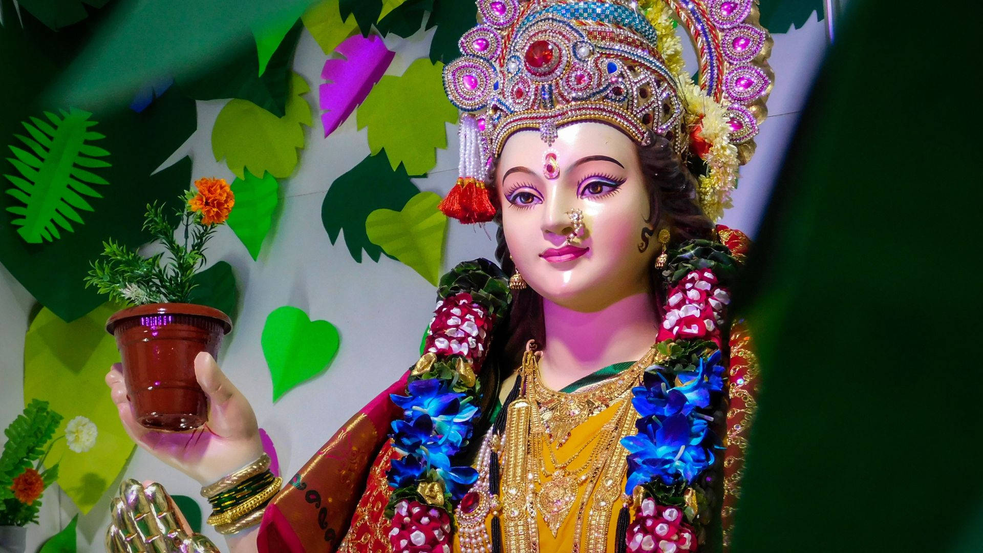 durga mata HD 桌布 壁紙 — 一座色彩鮮豔的印度教女神雕像，飾有珠寶和鮮花，在色彩繽紛的樹葉背景下捧著一盆萬壽菊。