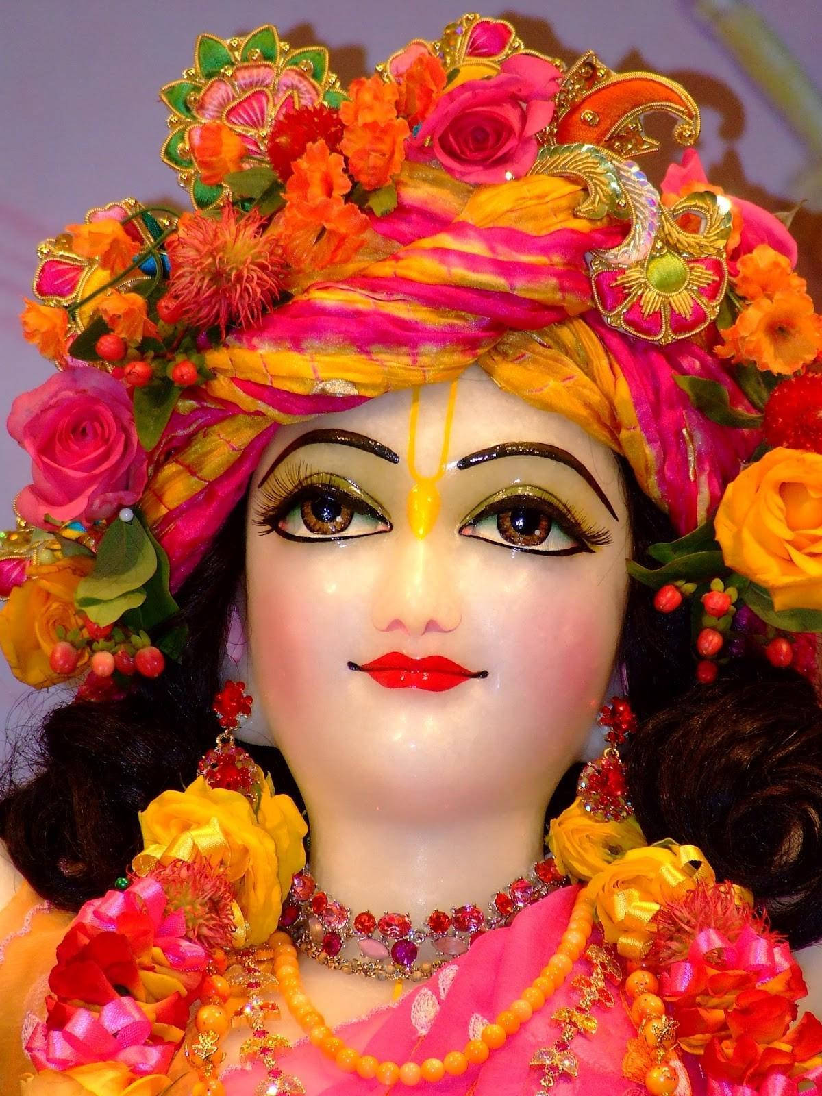 krishna ji 桌布 壁紙 — 一座色彩鮮豔的神像，裝飾著五顏六色的花朵、金色飾物和亮麗的粉紅色及黃色衣服。