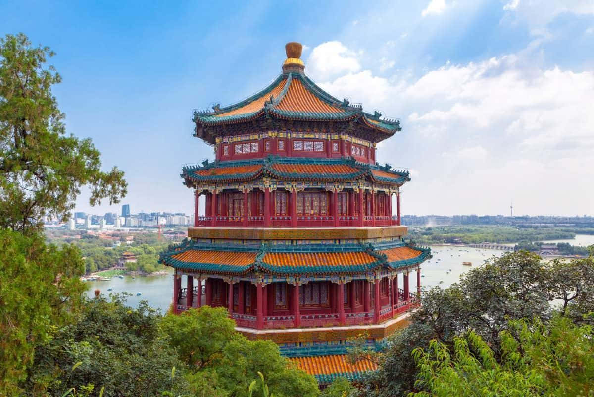 Blue Sky 桌布 壁紙 — 一座色彩鮮豔的紅橙色傳統中國寶塔，配以綠色瓦片屋頂，雄偉地矗立在山坡上，俯瞰著寧靜的湖泊和遠處的城市景觀，在明亮的藍天下。