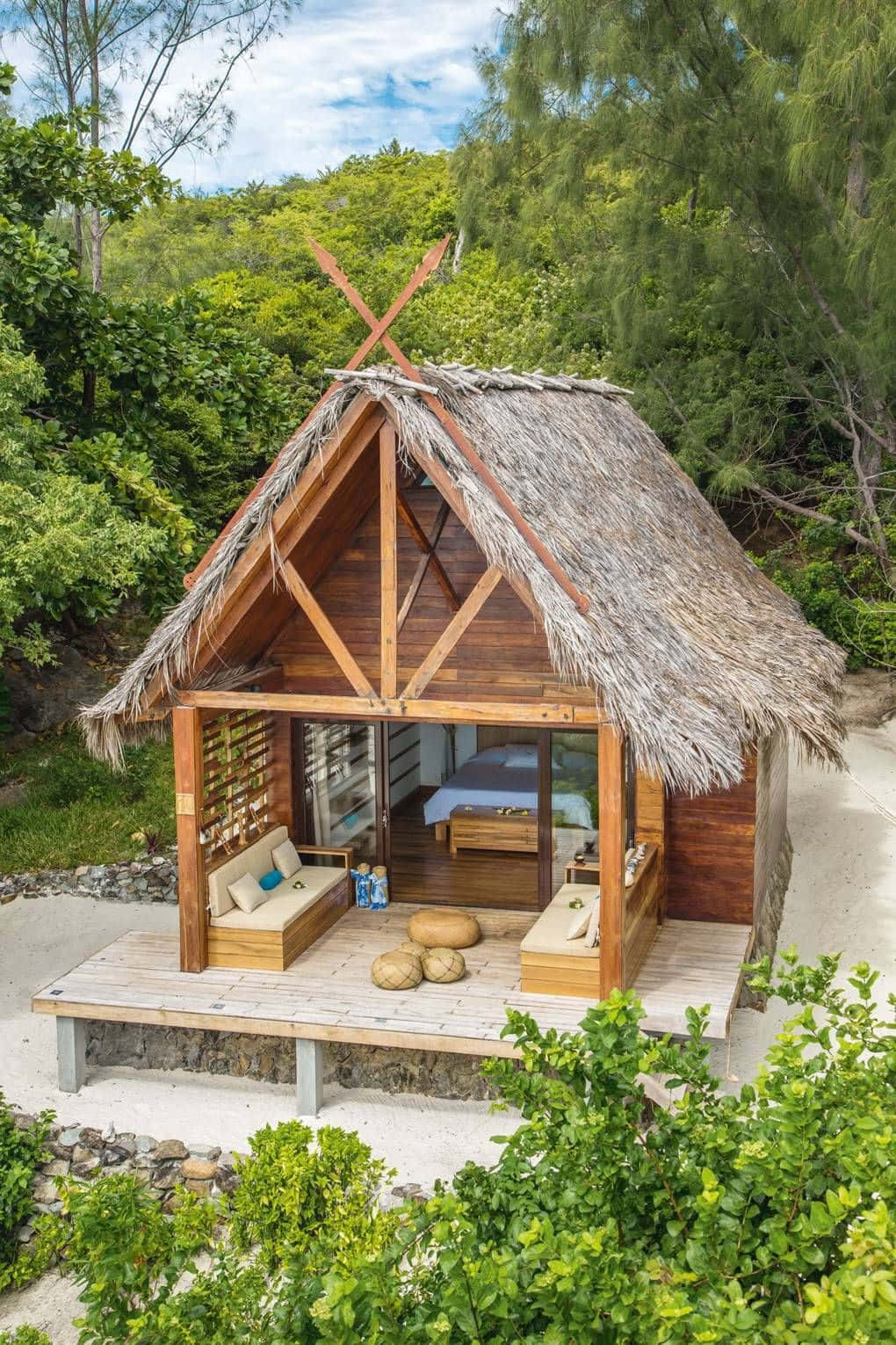 Cottage 桌布 壁紙 — 一座茅草屋頂的木製小屋坐落在沙灘上，周圍環繞著茂密的綠樹，頭頂是明亮的藍天。