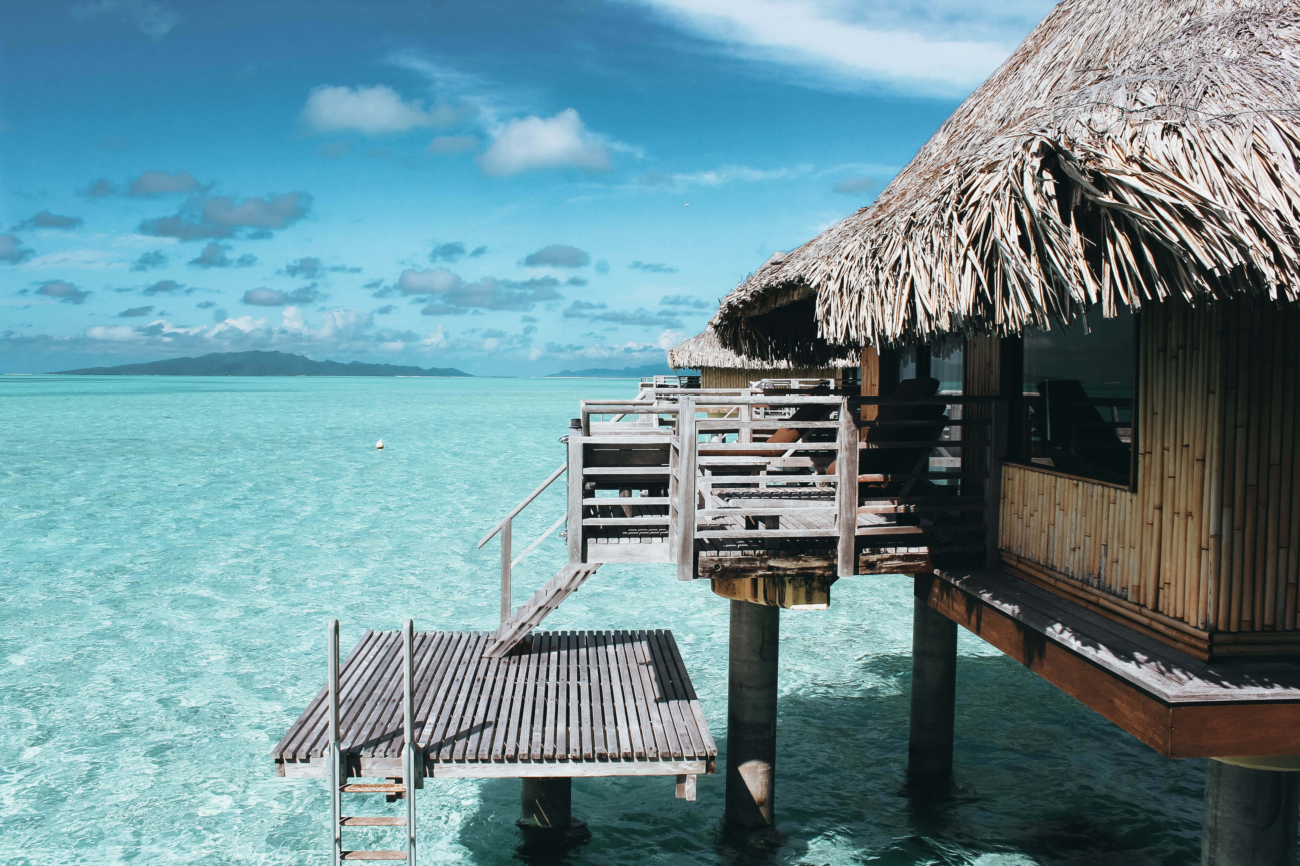 Beach Huts 桌布 壁紙 — 一座茅草屋頂的木製水上平房矗立在碧綠海水之上的支柱上，上方是明亮的藍天，點綴著幾朵雲。