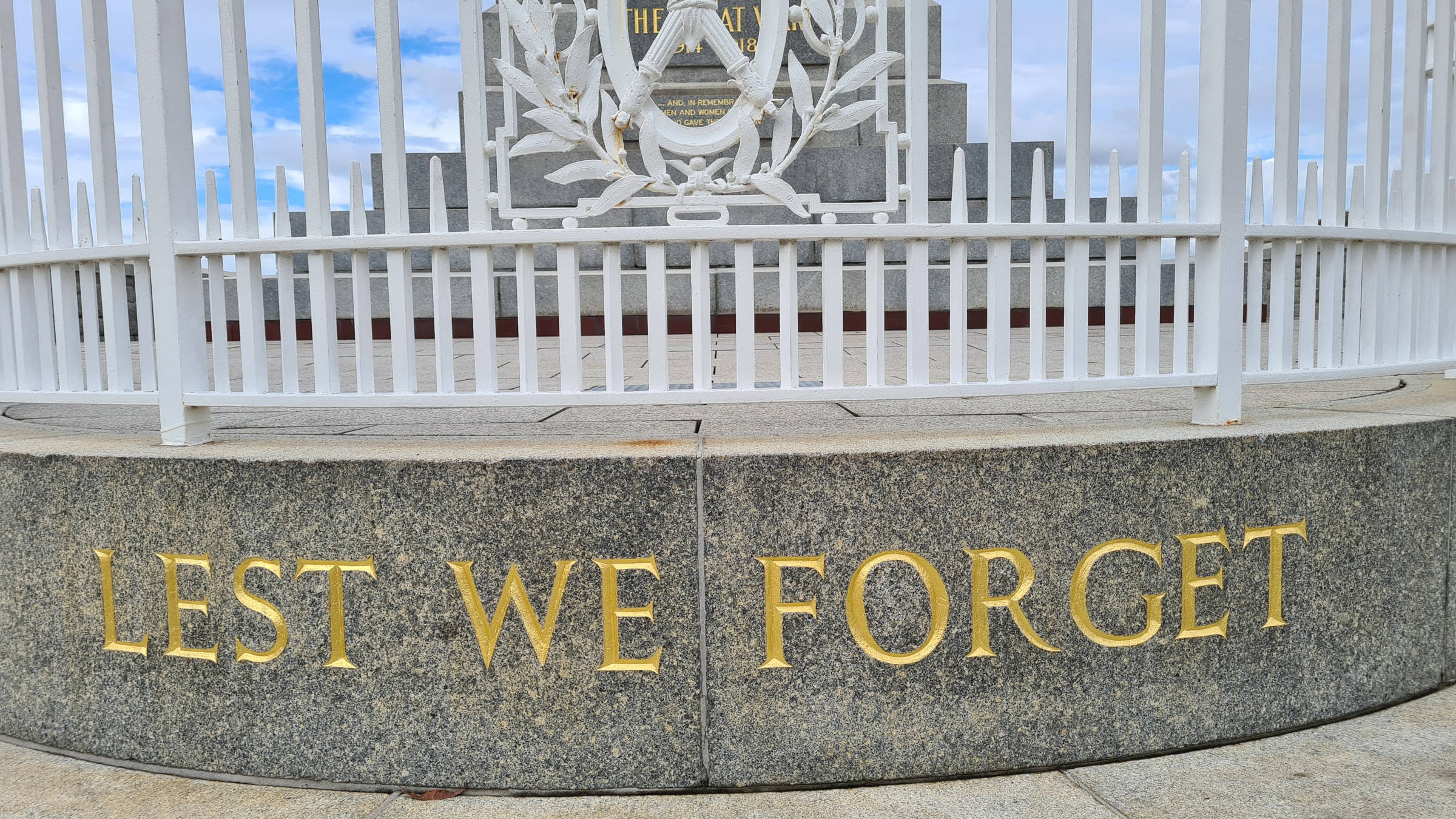 Remembrance 桌布 壁紙 — 一座莊嚴的戰爭紀念碑，花崗岩上刻有金色「LEST WE FORGET」字樣，在部分多雲的天空下以白色尖樁籬笆圍繞。