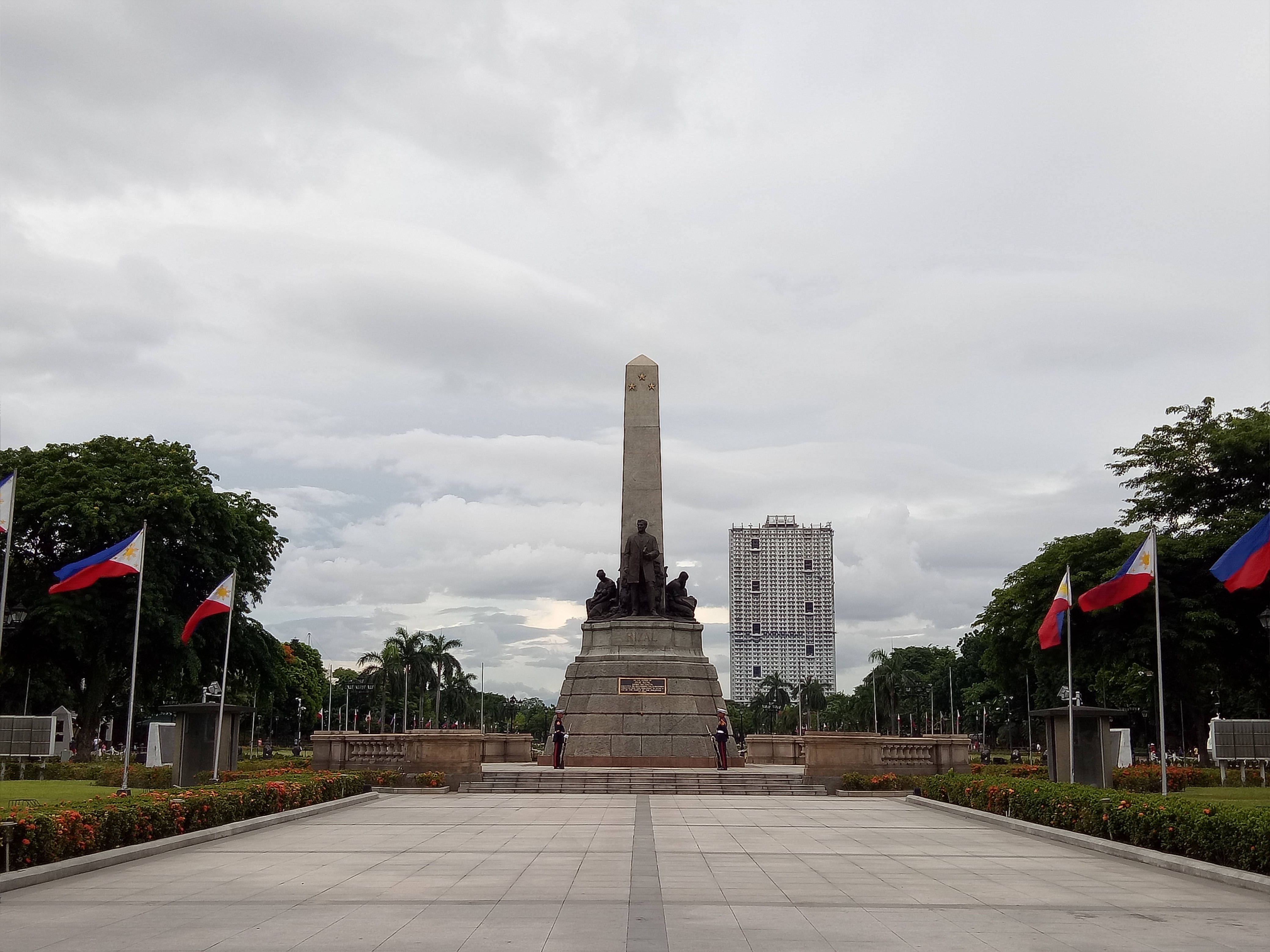Manila 桌布 壁紙 — 一座莊嚴的紀念碑，上有青銅雕像，矗立在寬闊廣場中央，兩旁菲律賓國旗飄揚，天空多雲。