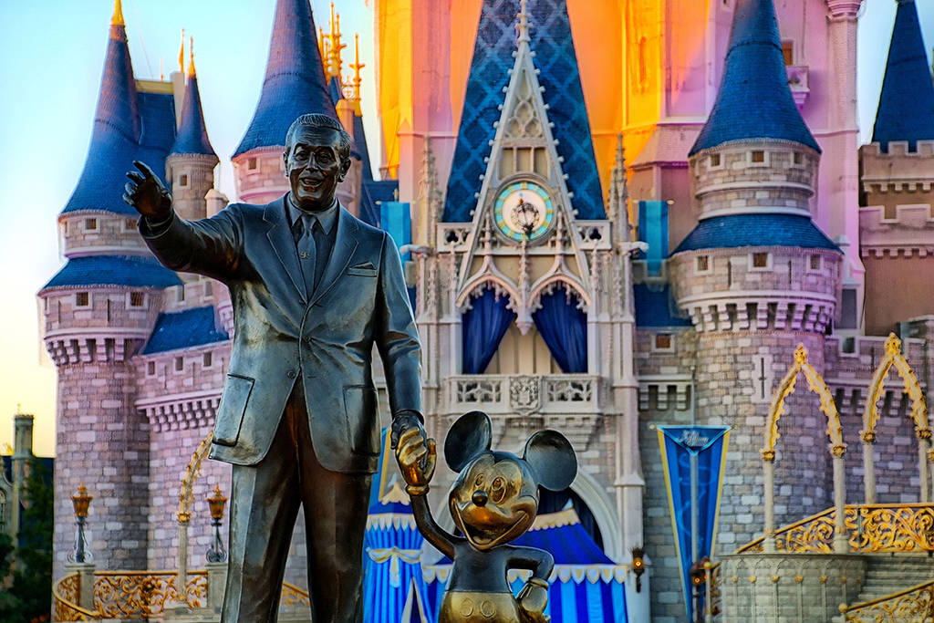 Disney Castle 桌布 壁紙 — 一座華特迪士尼抱著米奇老鼠的銅像，矗立在沐浴於金色夕陽光芒下的標誌性灰姑娘城堡前。