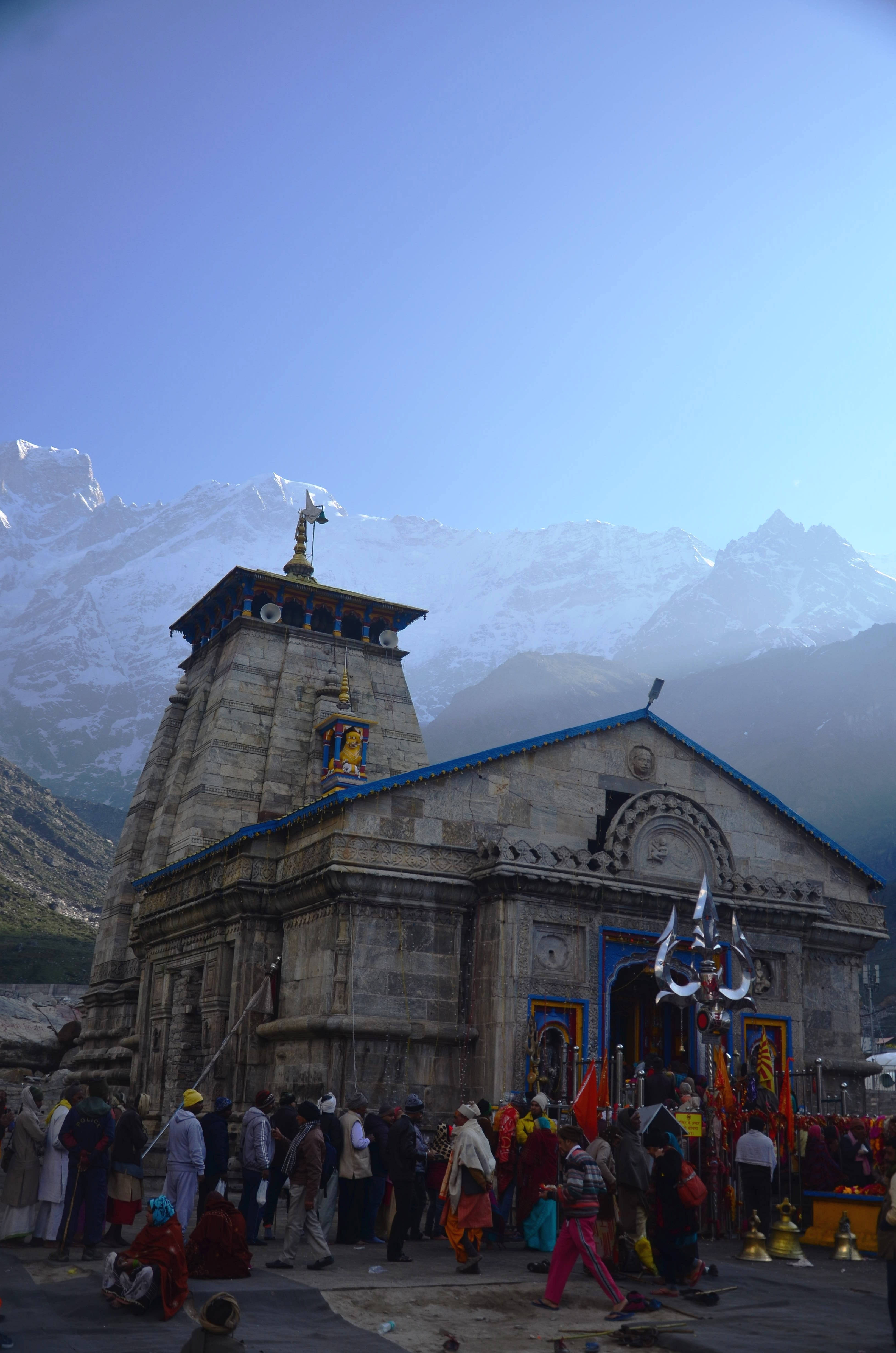 Kedarnath 桌布 壁紙 — 一座藍色屋頂的石廟矗立在積雪的山峰前，朝聖者在晴朗的藍天下聚集在入口處。