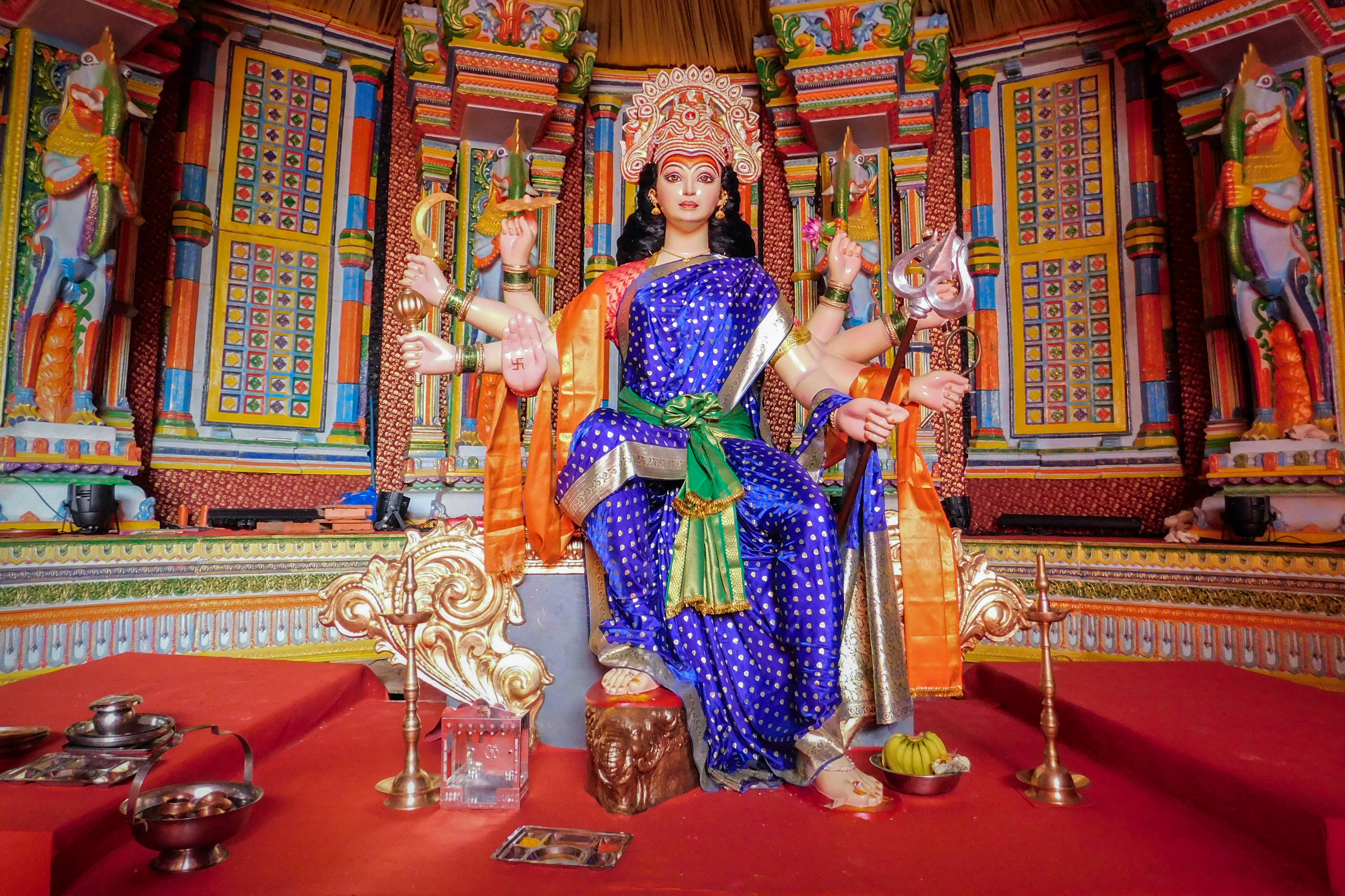 maa durga 桌布 壁紙 — 一座藍金色、色彩鮮豔的多臂女神雕像，威嚴地坐在紅色基座上，位於裝飾著彩色圖案和供品的華麗寺廟內。