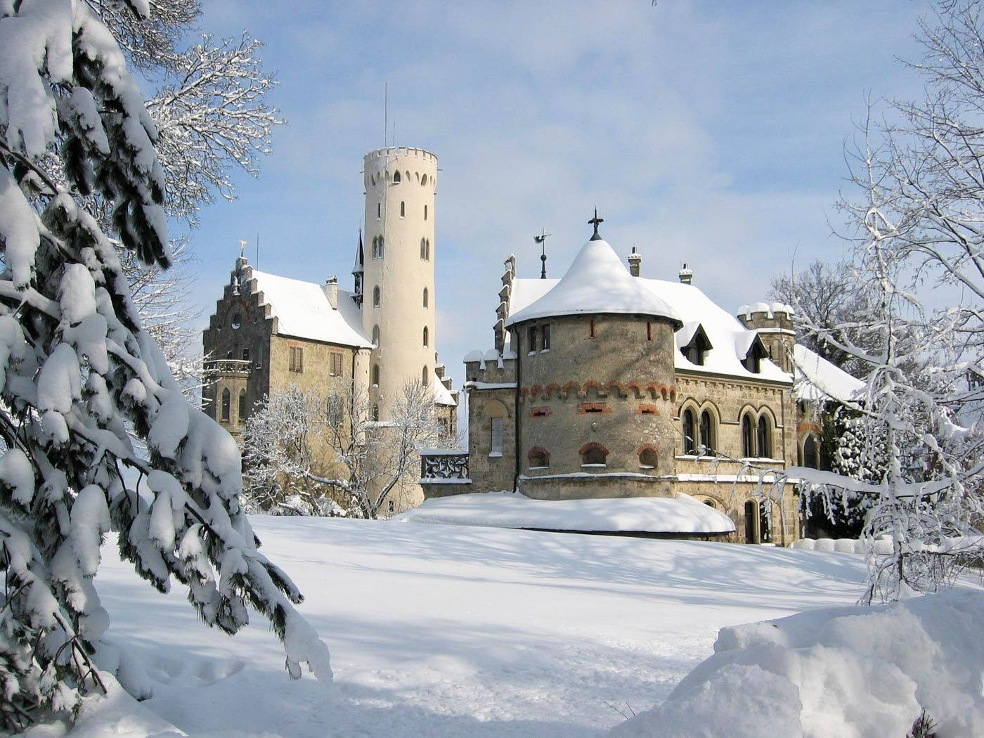 Liechtenstein 桌布 壁紙 — 一座被雪覆蓋的中世紀城堡，擁有石塔和拱形窗戶，在晴朗的藍天下靜靜矗立，被積雪的常青樹枝環繞。