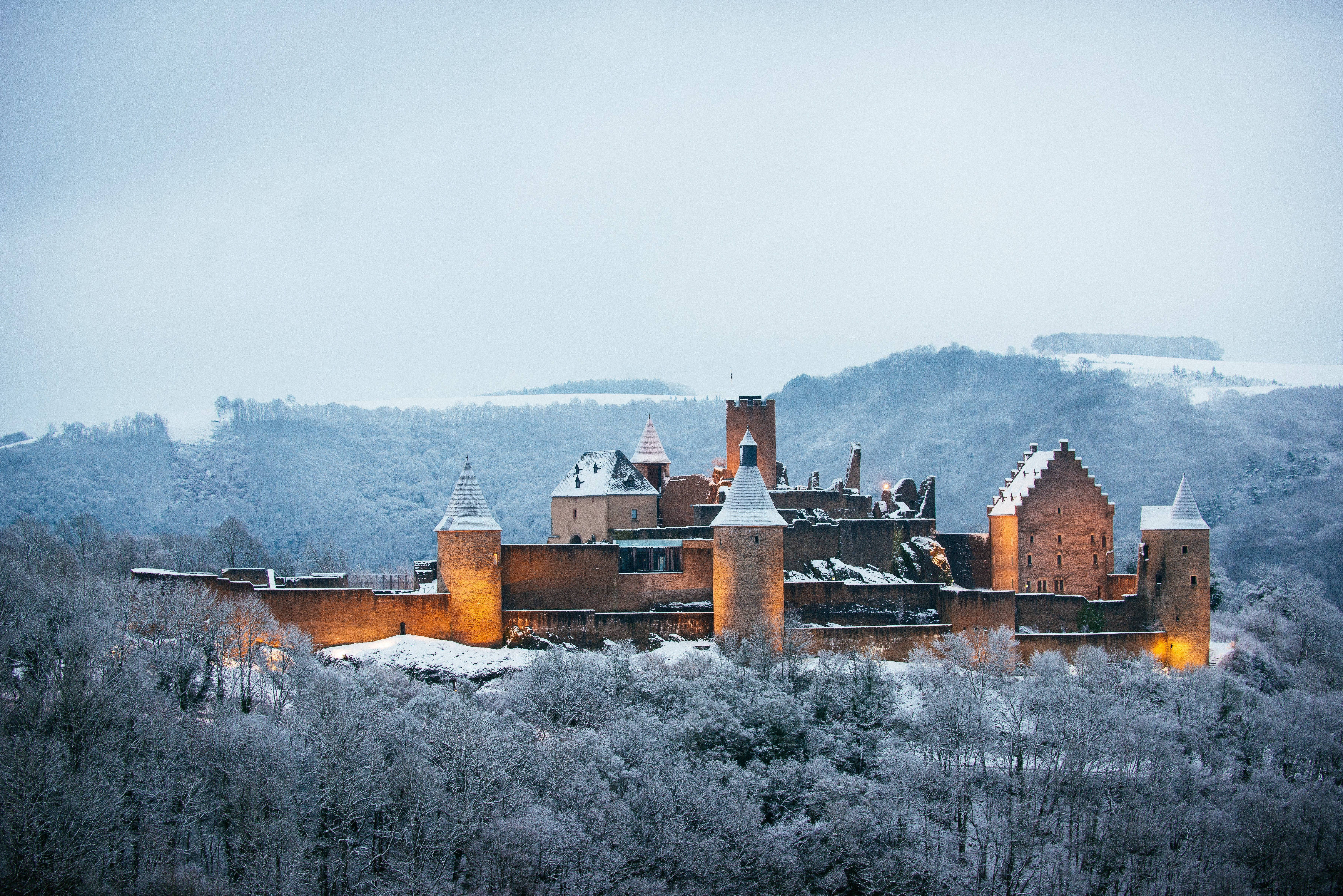 Luxembourg 桌布 壁紙 — 一座被雪覆蓋的中世紀城堡，溫暖發光的窗戶，在蒼白的冬日天空下，雄偉地矗立在森林覆蓋的山丘上。