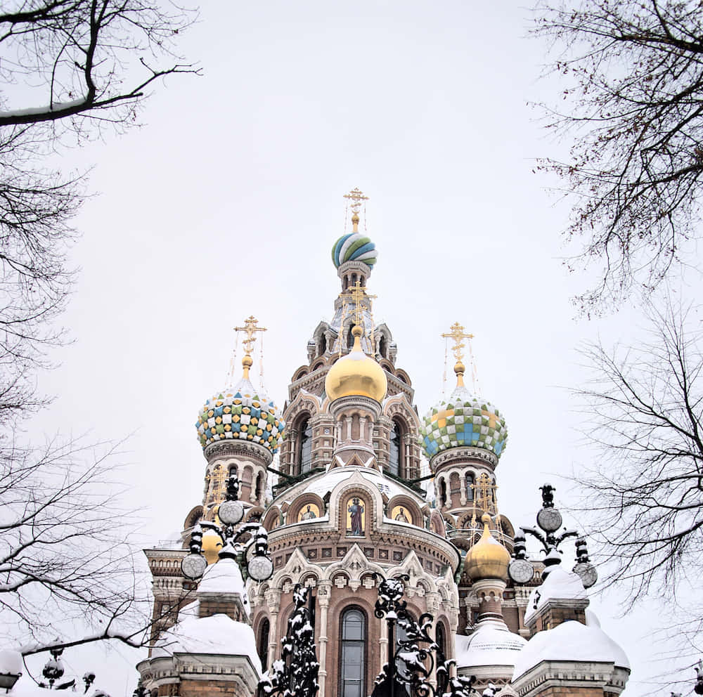 Beautiful Architecture 桌布 壁紙 — 一座被雪覆蓋的俄羅斯東正教教堂，擁有色彩繽紛的洋蔥形圓頂和金色十字架，在光禿禿的樹枝框住的蒼白冬日天空下巍然聳立。