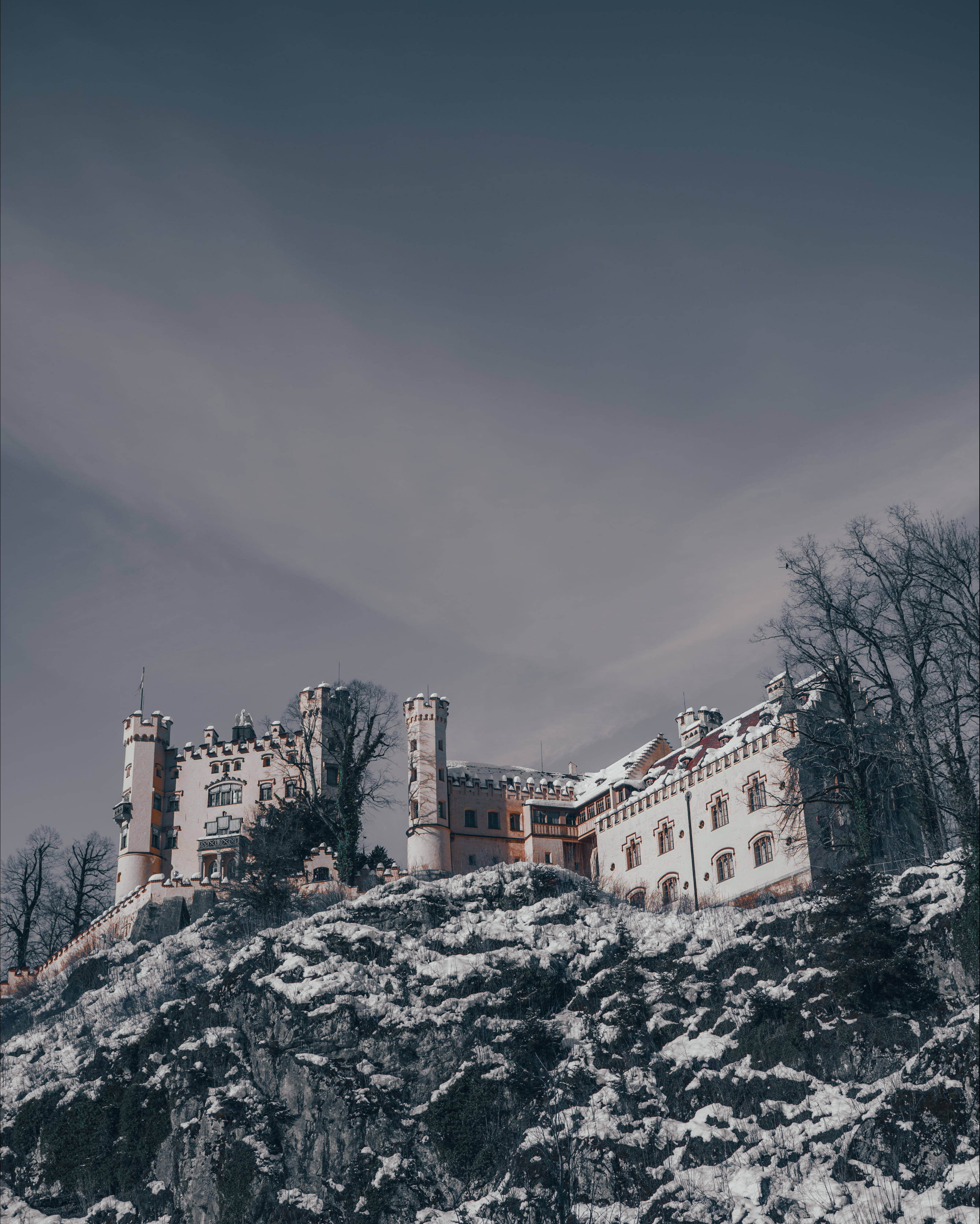 Frozen Castle 桌布 壁紙 — 一座被雪覆蓋的城堡坐落在岩石山頂上，天空陰沉灰暗，樹木光禿。