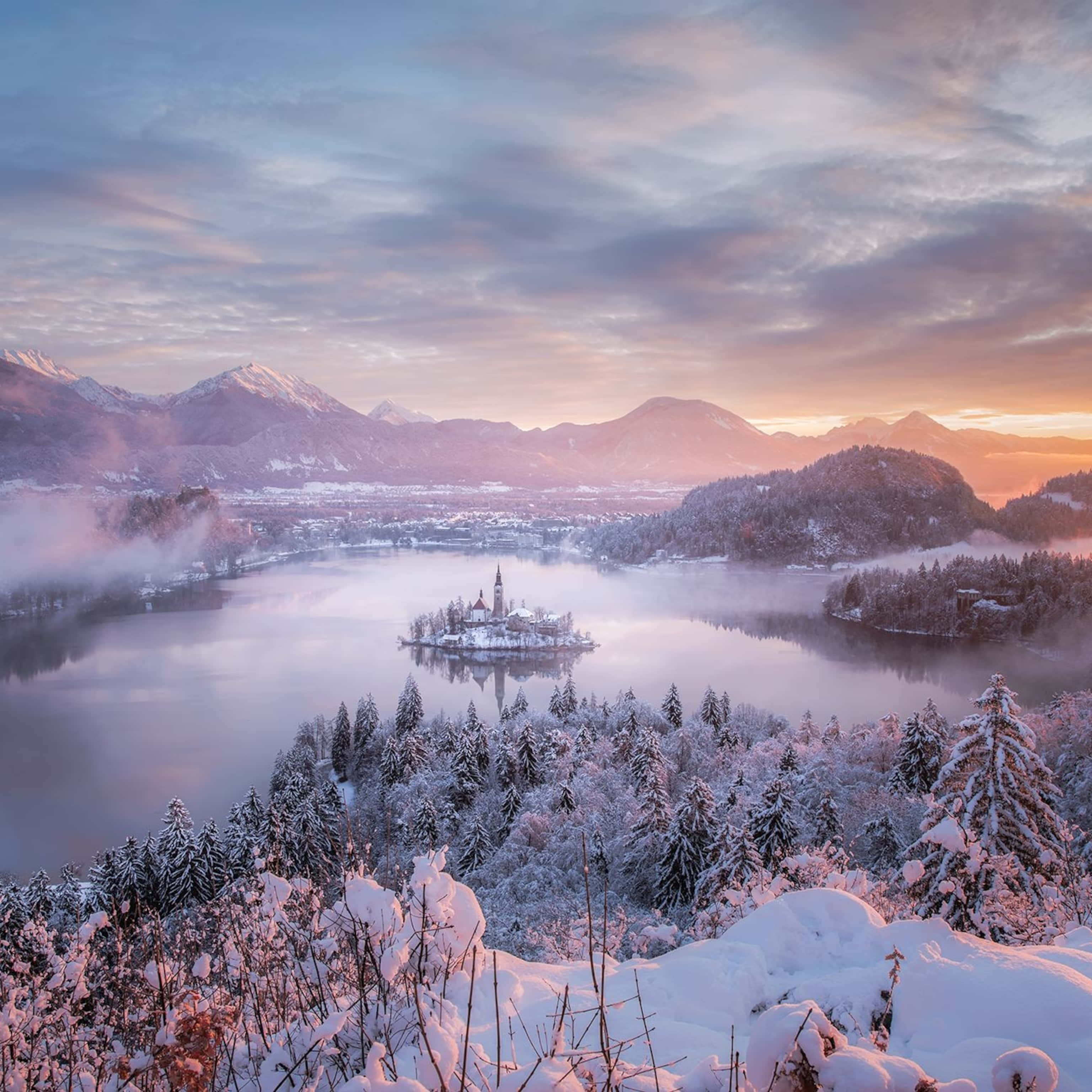 Landscapes 桌布 壁紙 — 一座被雪覆蓋的島嶼教堂在日出時分，在霧氣瀰漫的湖面上閃耀，周圍是森林覆蓋的山脈。
