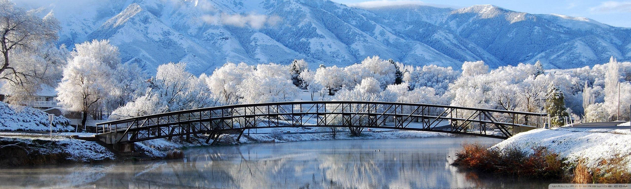 冬季 桌布 壁紙 — 一座被雪覆蓋的拱橋橫跨平靜的河流，背景中有結霜的樹木和雄偉的山脈。