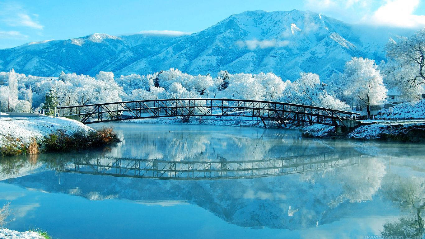 Lake 桌布 壁紙 — 一座被雪覆蓋的拱橋橫跨平靜的藍色河流，映照出白雪覆蓋的山脈和結霜的樹木，天空晴朗。