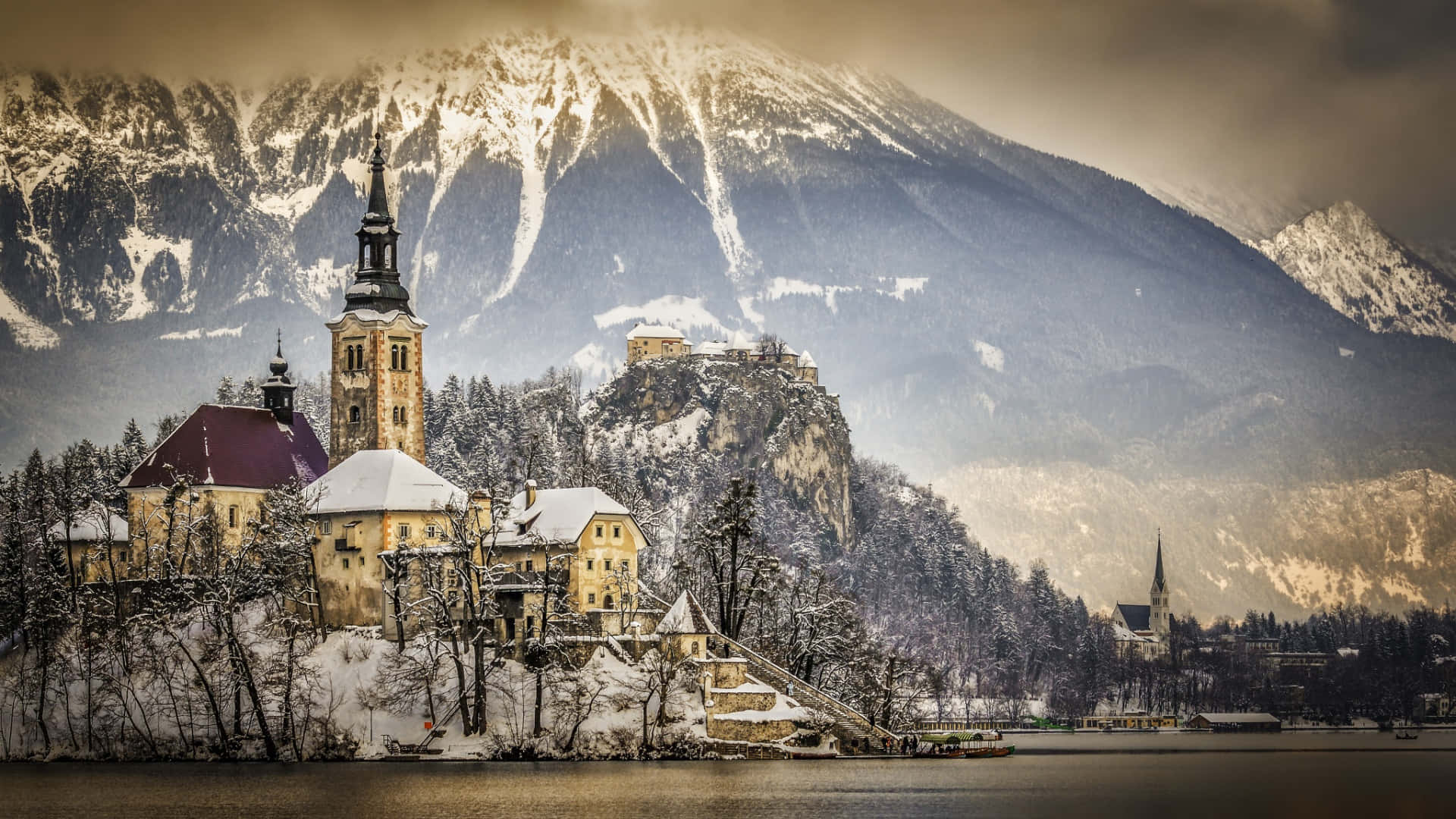 Alps 桌布 壁紙 — 一座被雪覆蓋的教堂和城堡坐落在湖邊懸崖上，背景是陰沉天空下高聳的山峰。