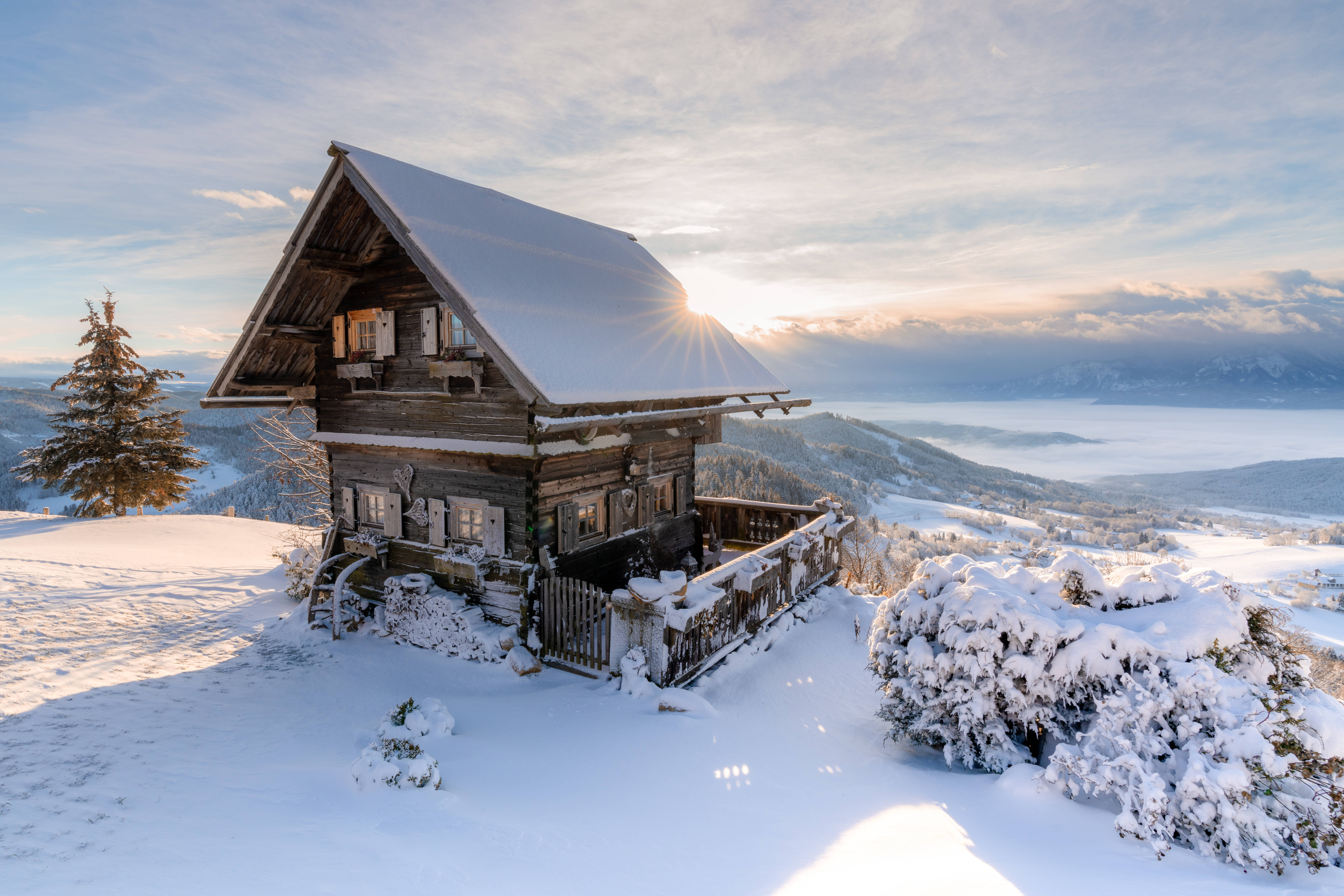 Austria 桌布 壁紙 — 一座被雪覆蓋的木屋坐落在山脊上，沐浴在金色的日出光芒中，下方是遙遠的山峰和山谷。