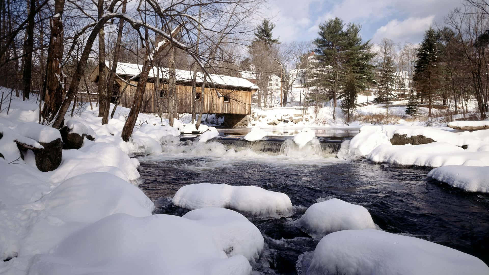 冬天 桌布 壁紙 — 一座被雪覆蓋的木橋橫跨湍急的河流，河中有結霜的岩石和光禿的樹木，在明亮的冬日天空下。