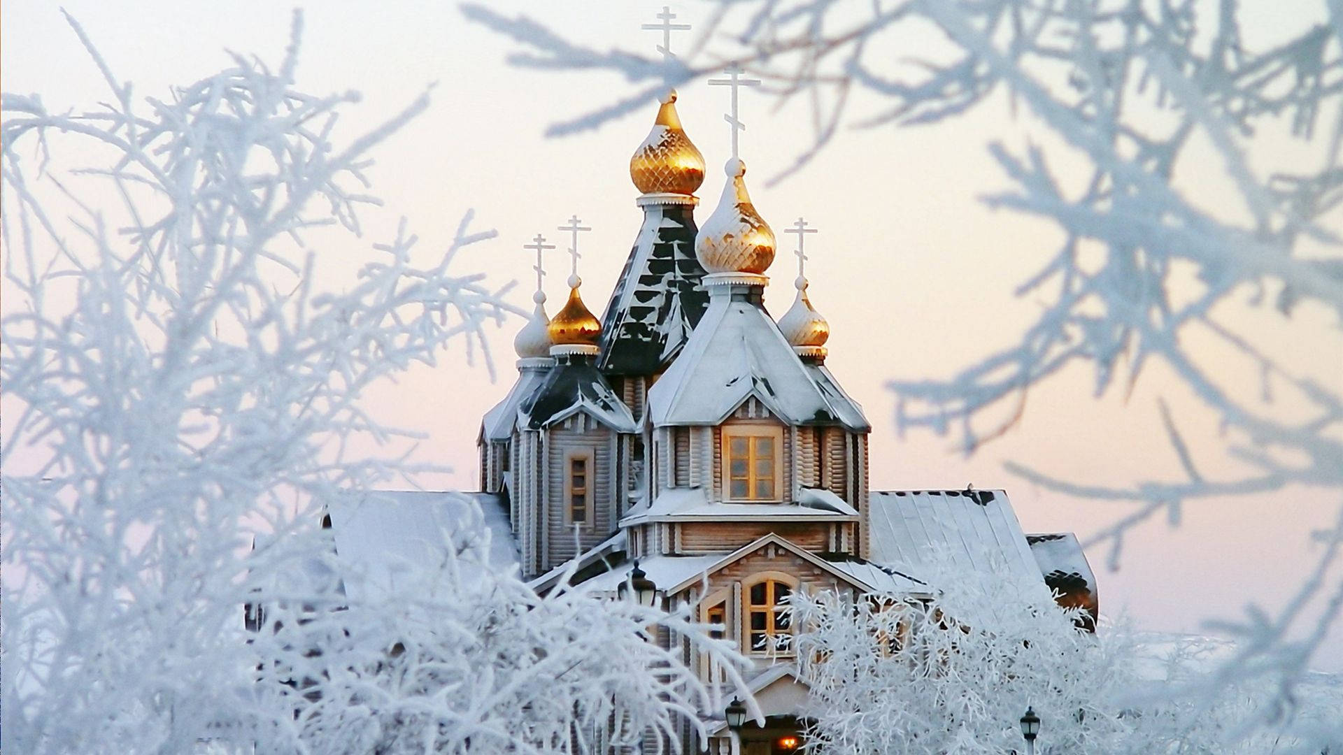 Russia 桌布 壁紙 — 一座被雪覆蓋的木製教堂，金色圓頂在冬日黃昏透過結霜的樹枝溫暖地發光。