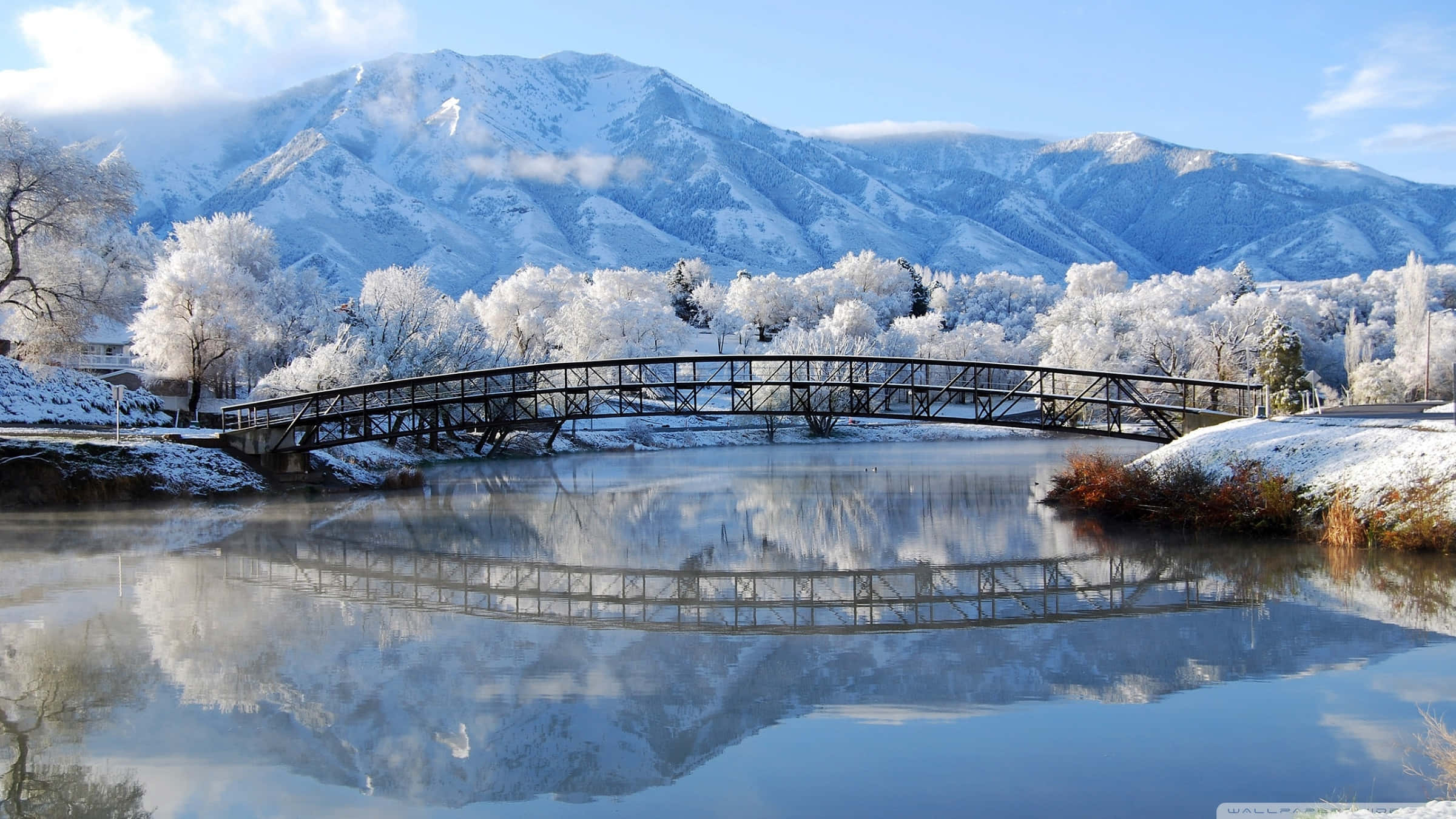 Beautiful Winter 桌布 壁紙 — 一座被雪覆蓋的橋樑拱起在平靜的河面上，映照著白雪覆蓋的山脈和結霜的樹木，在清澈的藍天下。