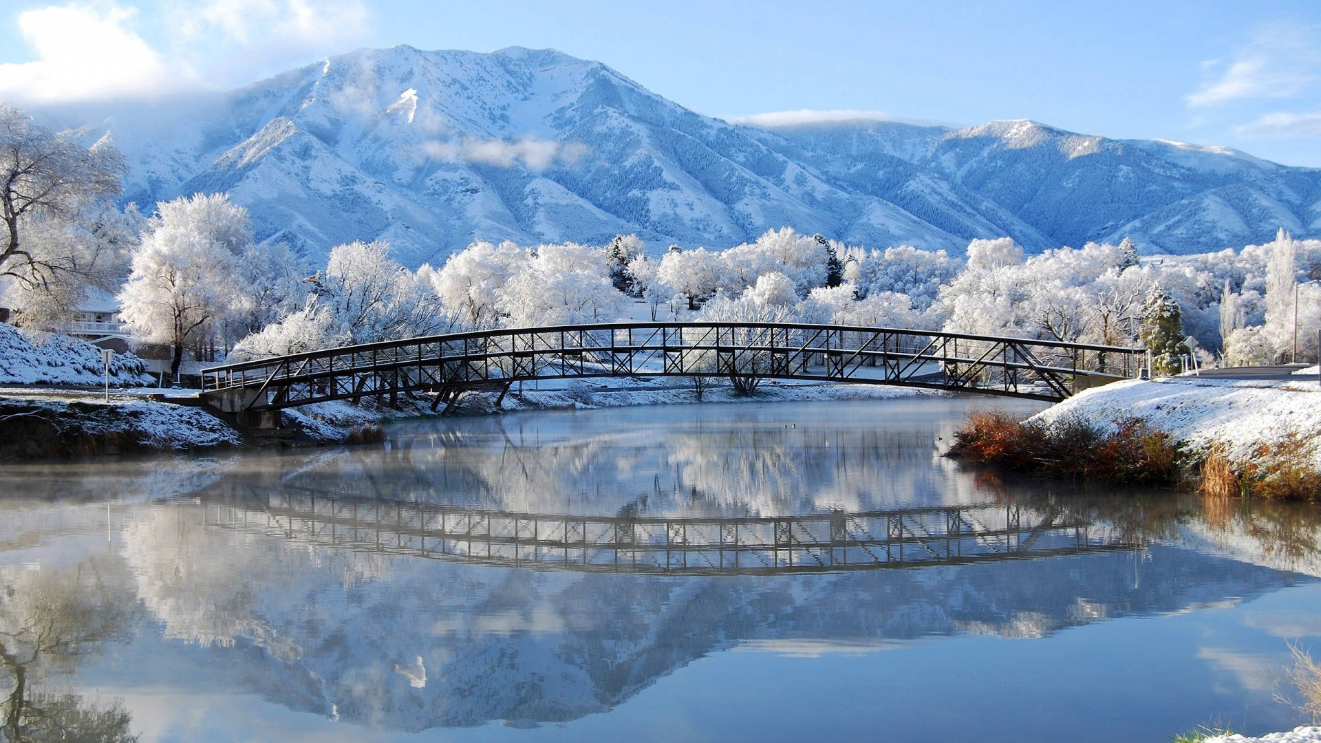 Winter Landscape 桌布 壁紙 — 一座被雪覆蓋的橋樑拱起在平靜的河面上，倒映著白雪皚皚的山脈和結霜的樹木，天空清澈湛藍。