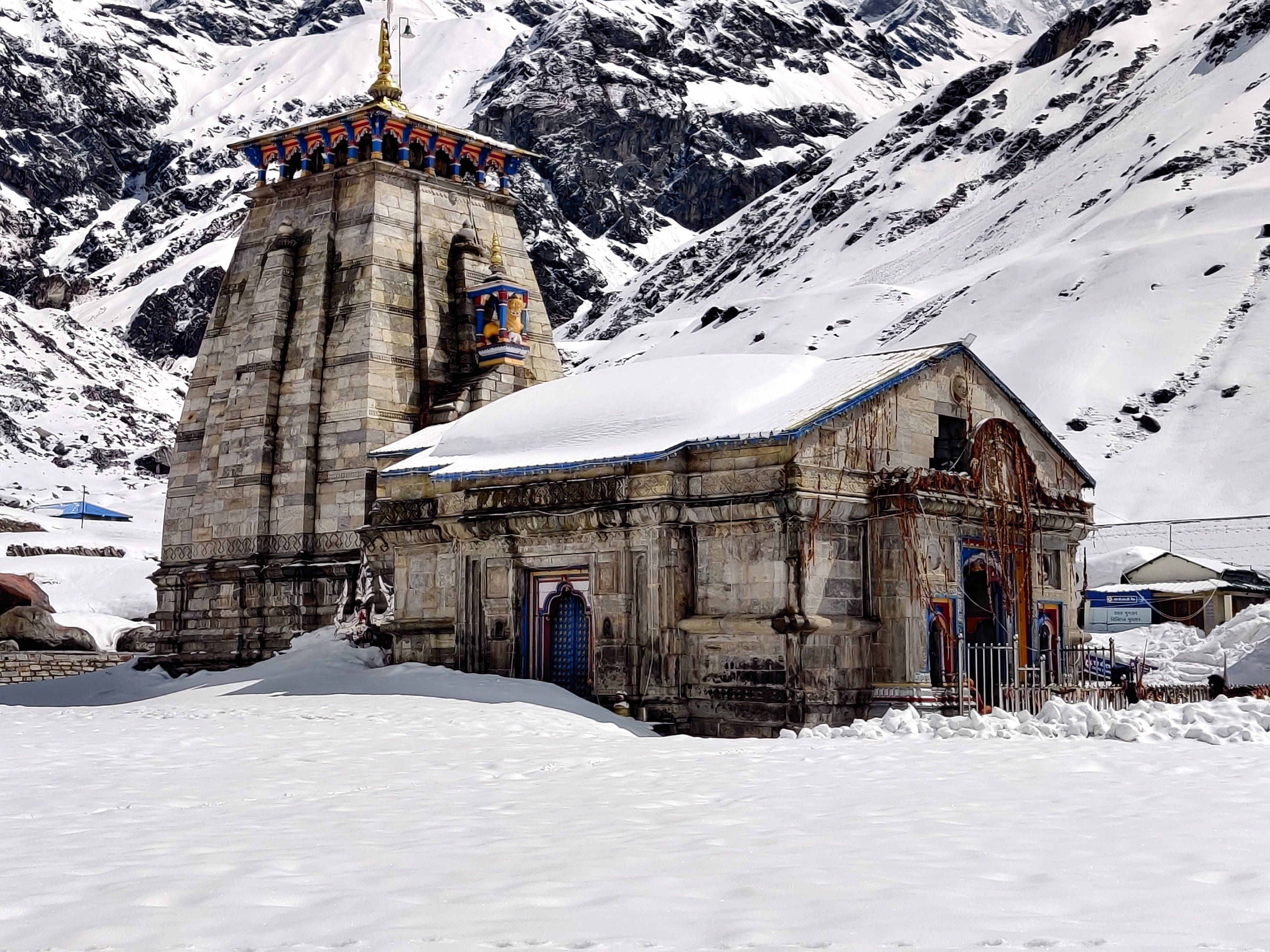 Kedarnath 桌布 壁紙 — 一座被雪覆蓋的石廟，帶有色彩繽紛的點綴，莊嚴地矗立在崎嶇、雪塵覆蓋的喜馬拉雅山峰背景下。