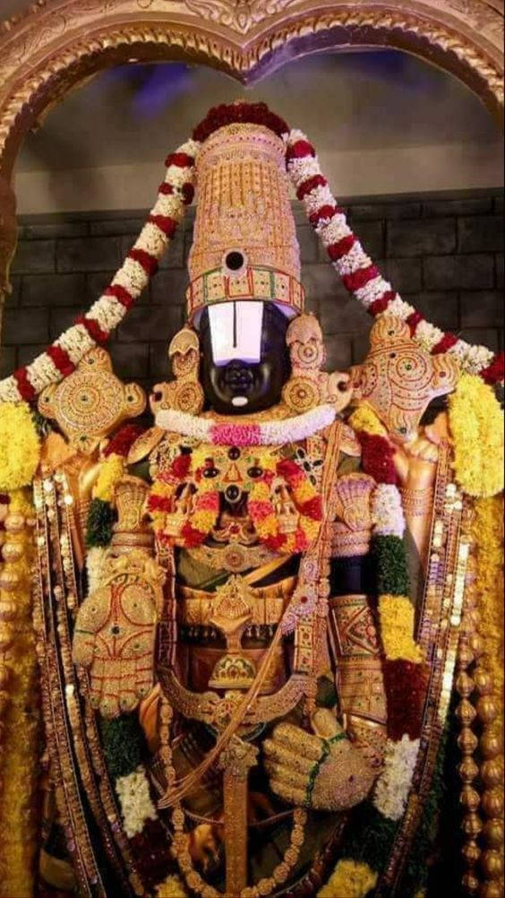 Lord Perumal 桌布 壁紙 — 一座裝飾華麗的黑色神像，披掛著鮮豔的花環和金飾，位於華麗的拱門下。