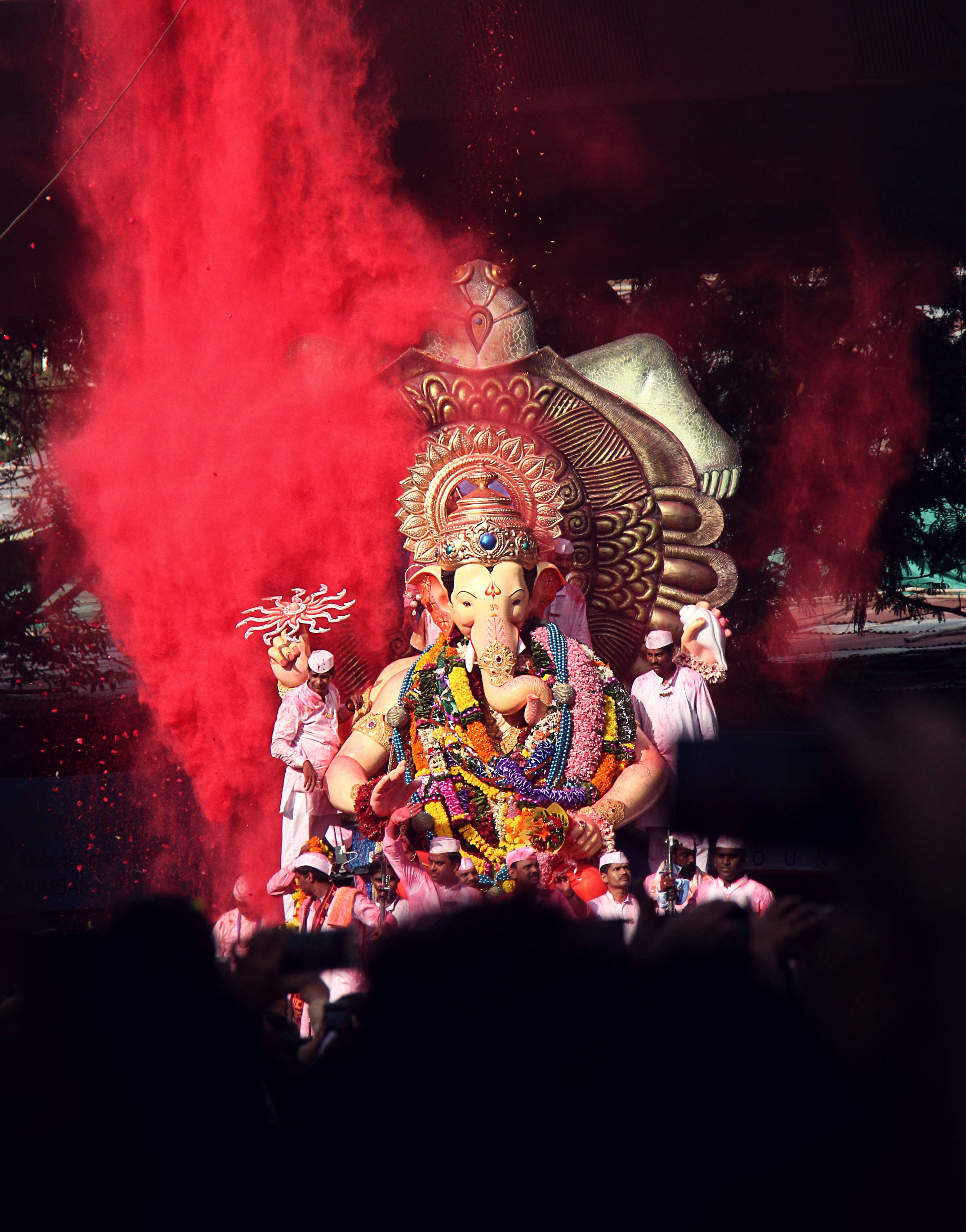 Ganesh Chaturthi 桌布 壁紙 — 一座裝飾著彩色花環的大型照明象神雕像，在節日遊行中矗立在紅色粉末的爆發中。