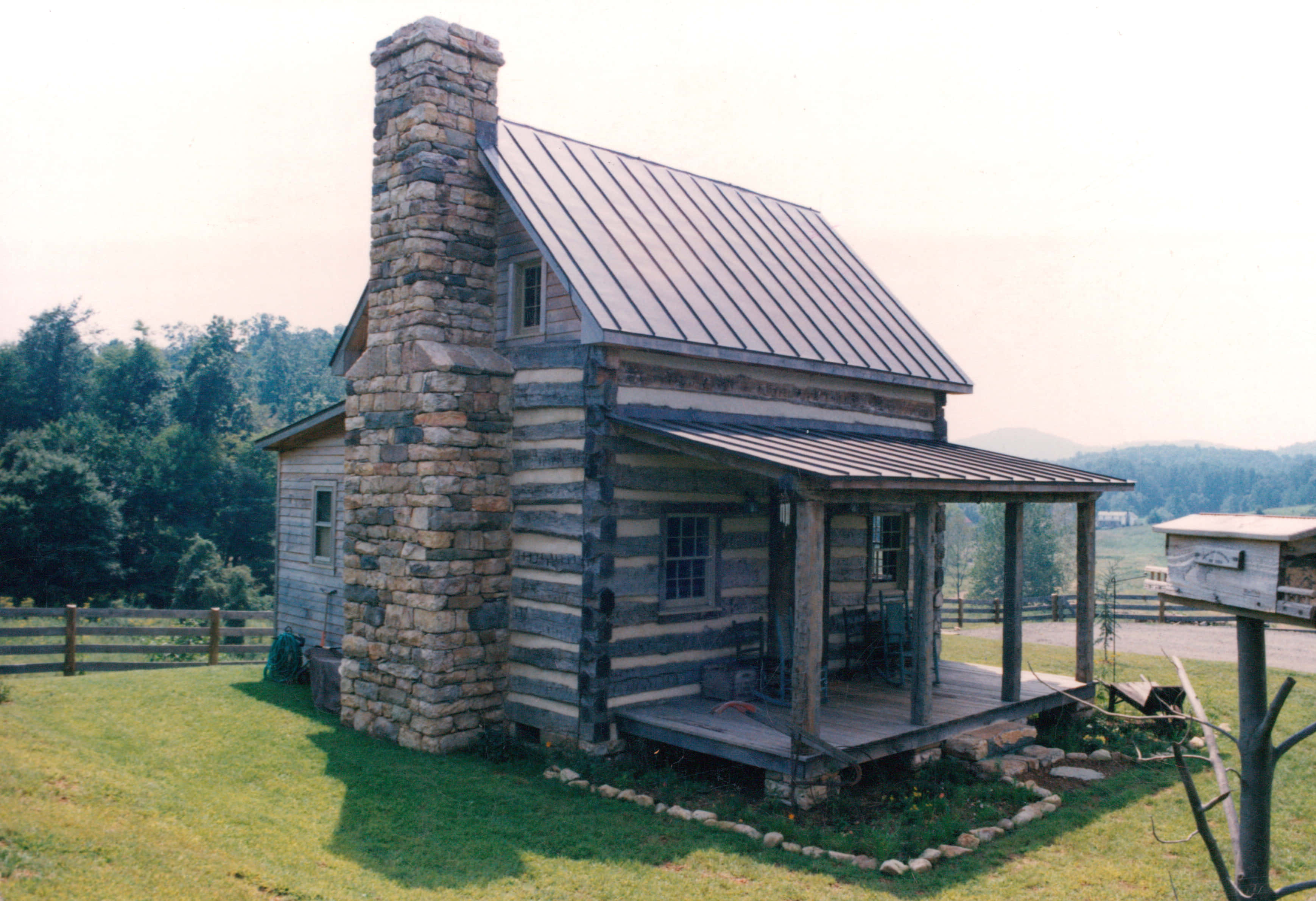 Log Cabin 桌布 壁紙 — 一座質樸的原木小屋，帶有石頭煙囪和金屬屋頂，坐落在被樹木和木柵欄環繞的草坡上。