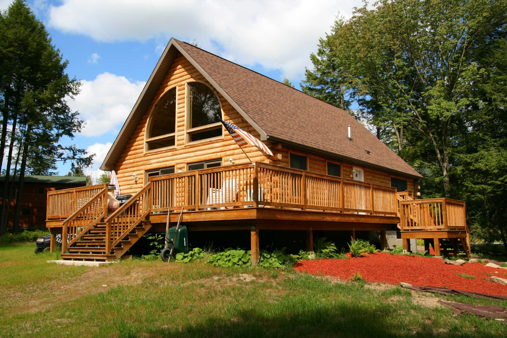 Log Cabin 桌布 壁紙 — 一座質樸的木屋，帶有大型木甲板和美國國旗，周圍環繞著高大的綠樹，天空湛藍，白雲朵朵。