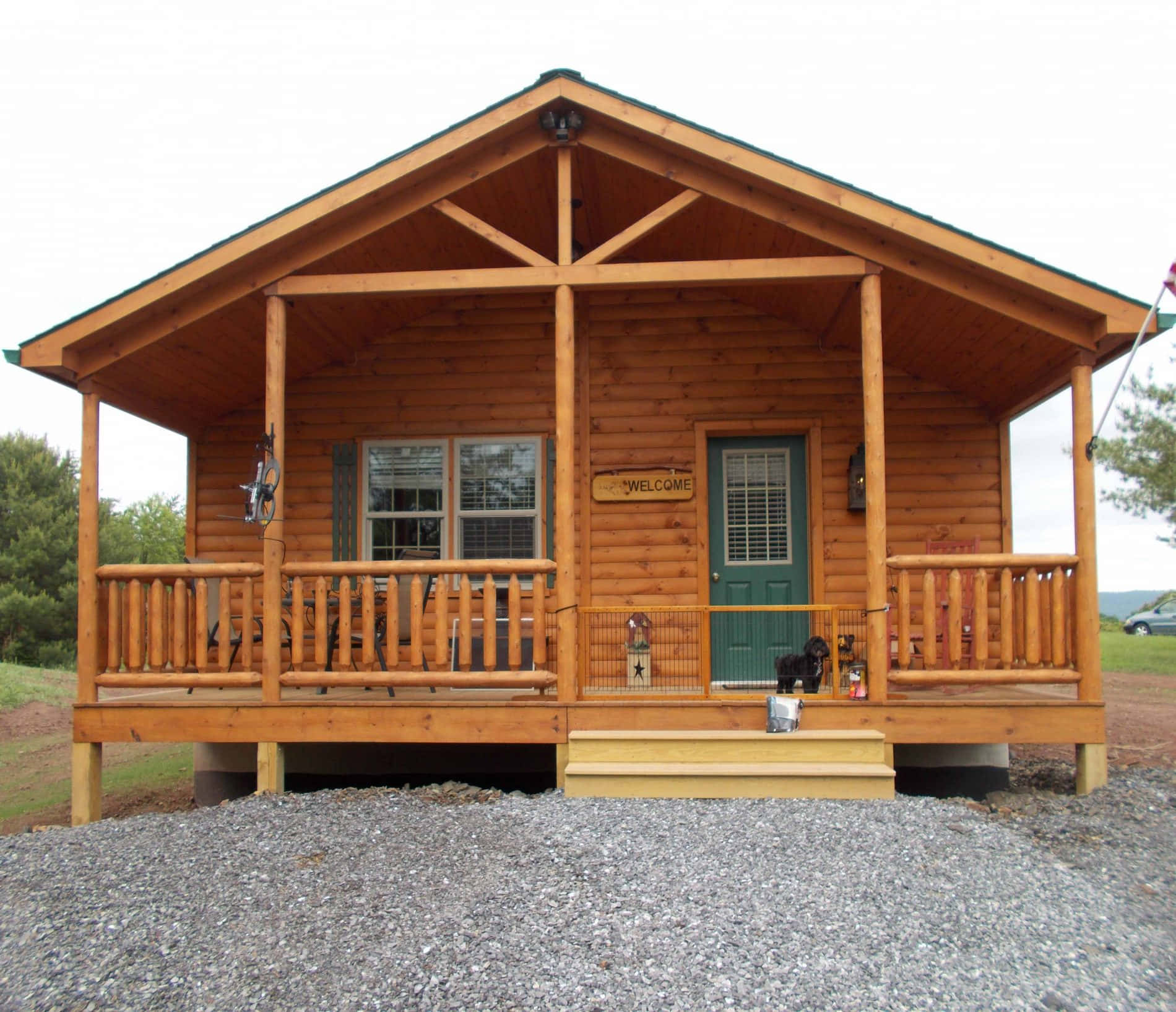 Log Cabin 桌布 壁紙 — 一座質樸的木屋，帶有有蓋門廊和綠色門，矗立在碎石地基上，周圍環繞著樹木。