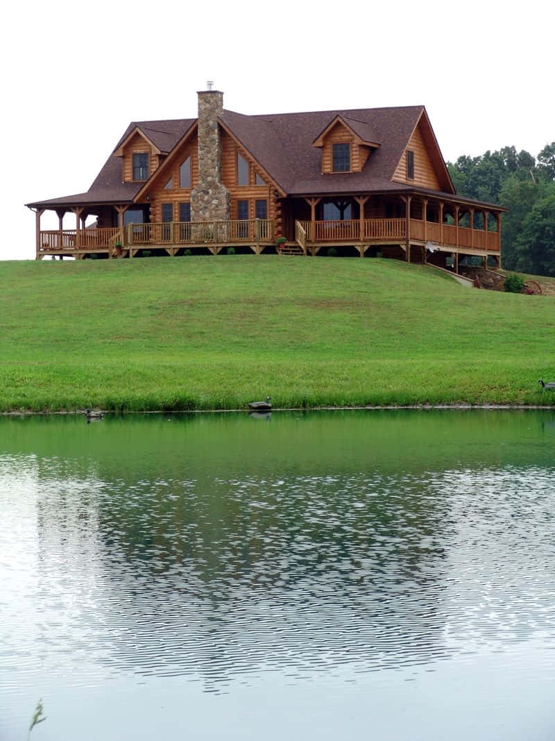 cabin 桌布 壁紙 — 一座質樸的木屋，帶有石頭煙囪，坐落在長滿青草的山丘上，俯瞰著平靜的湖泊，湖中有鴨子在游泳。