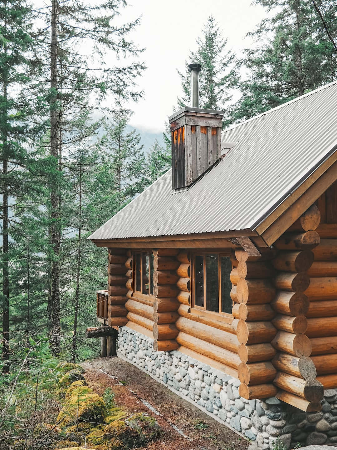 Log Cabin 桌布 壁紙 — 一座質樸的木屋，擁有石頭地基和金屬屋頂，坐落在霧氣瀰漫的森林中，周圍是高聳的常青樹。