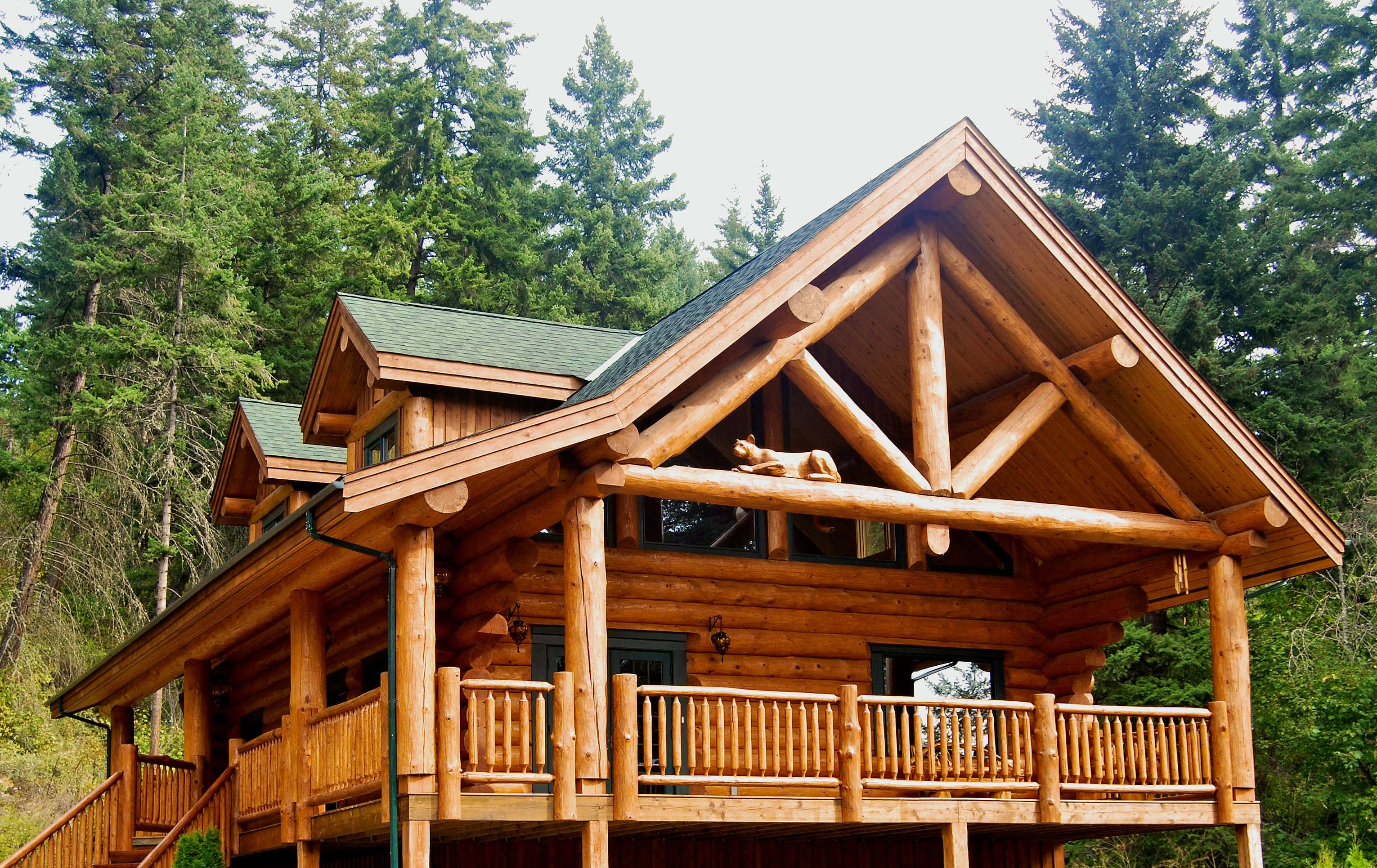 Log Cabin 桌布 壁紙 — 一座質樸的木屋，配有木欄杆和綠色屋頂，坐落在高大的常綠樹木之間。