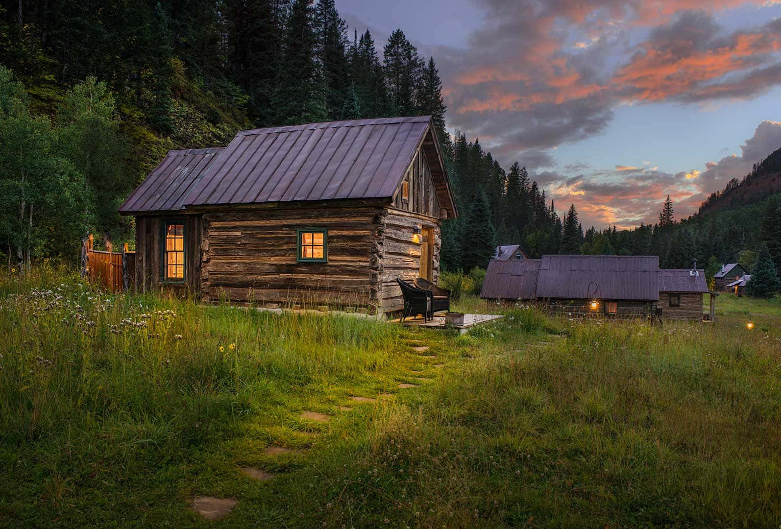 Log Cabin 桌布 壁紙 — 一座質樸的木屋在黃昏時分溫暖地發光，周圍是高高的草和松樹，天空呈現火熱的橙色和紫色。