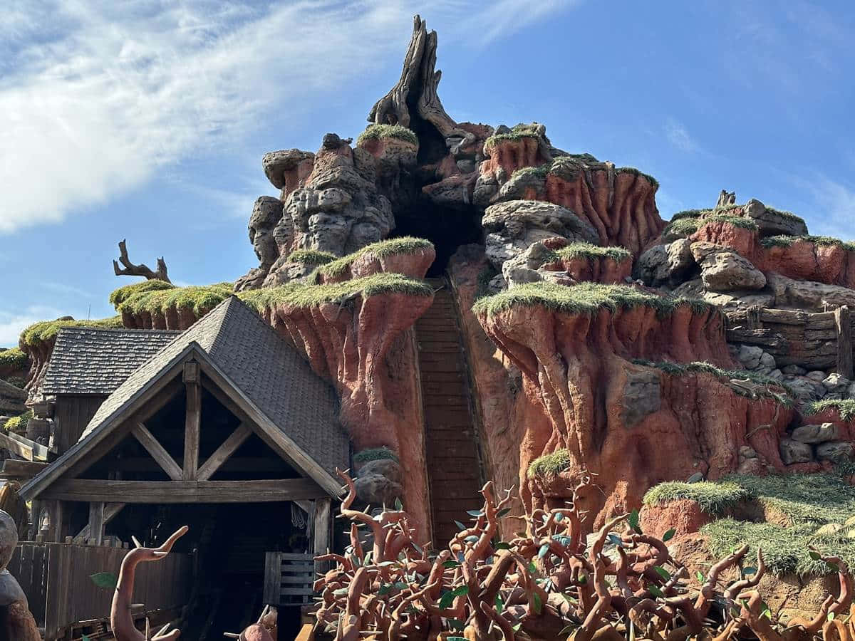 Splash Mountain 桌布 壁紙 — 一座質樸的木屋坐落在高聳、長滿苔蘚的紅色岩層腳下，上方是明亮的藍天。