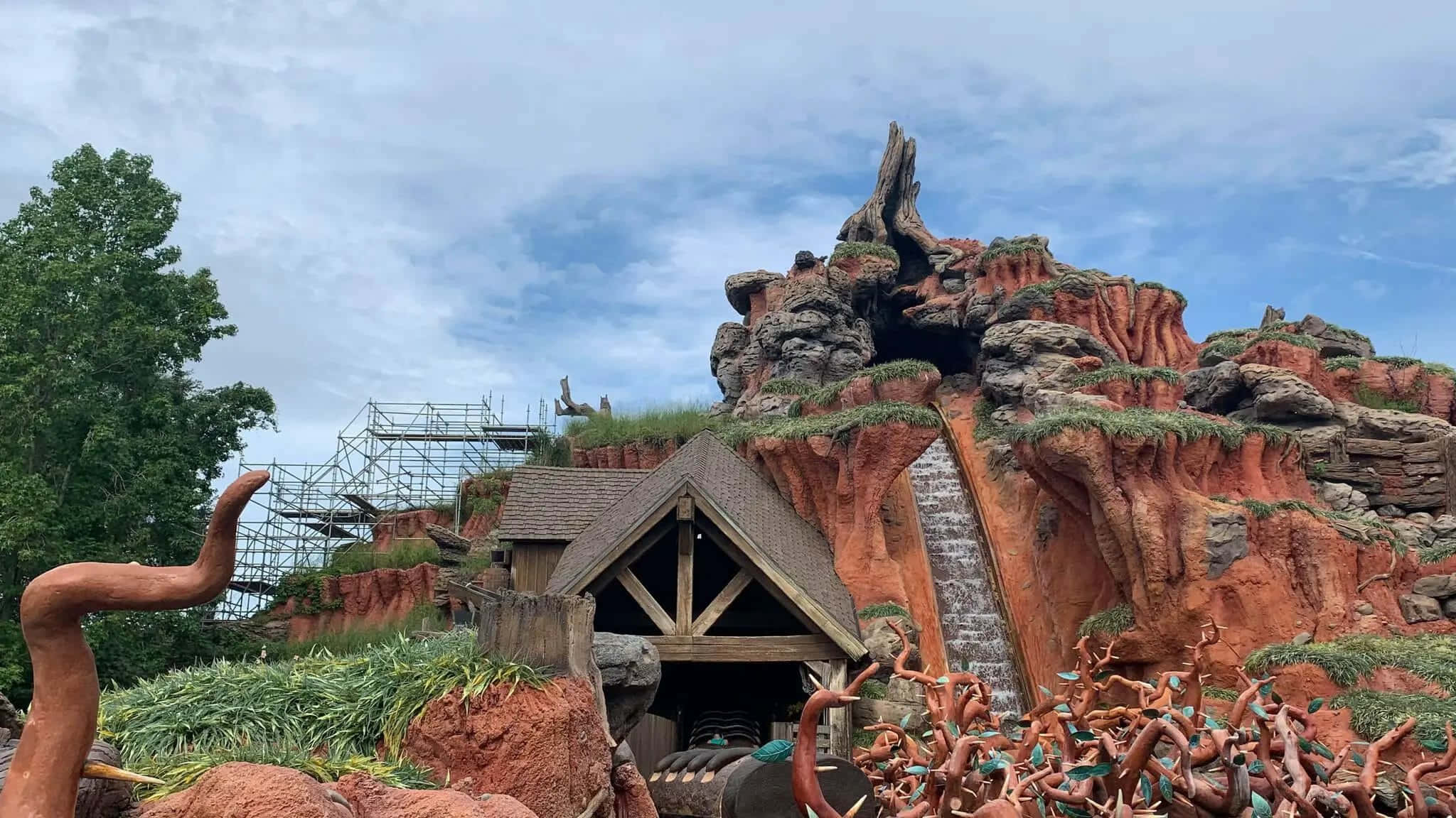 Splash Mountain 桌布 壁紙 — 一座質樸的木結構建築，坐落在崎嶇的紅色岩石峽谷中，有層疊瀑布和雕塑般的樹幹，天空部分多雲。