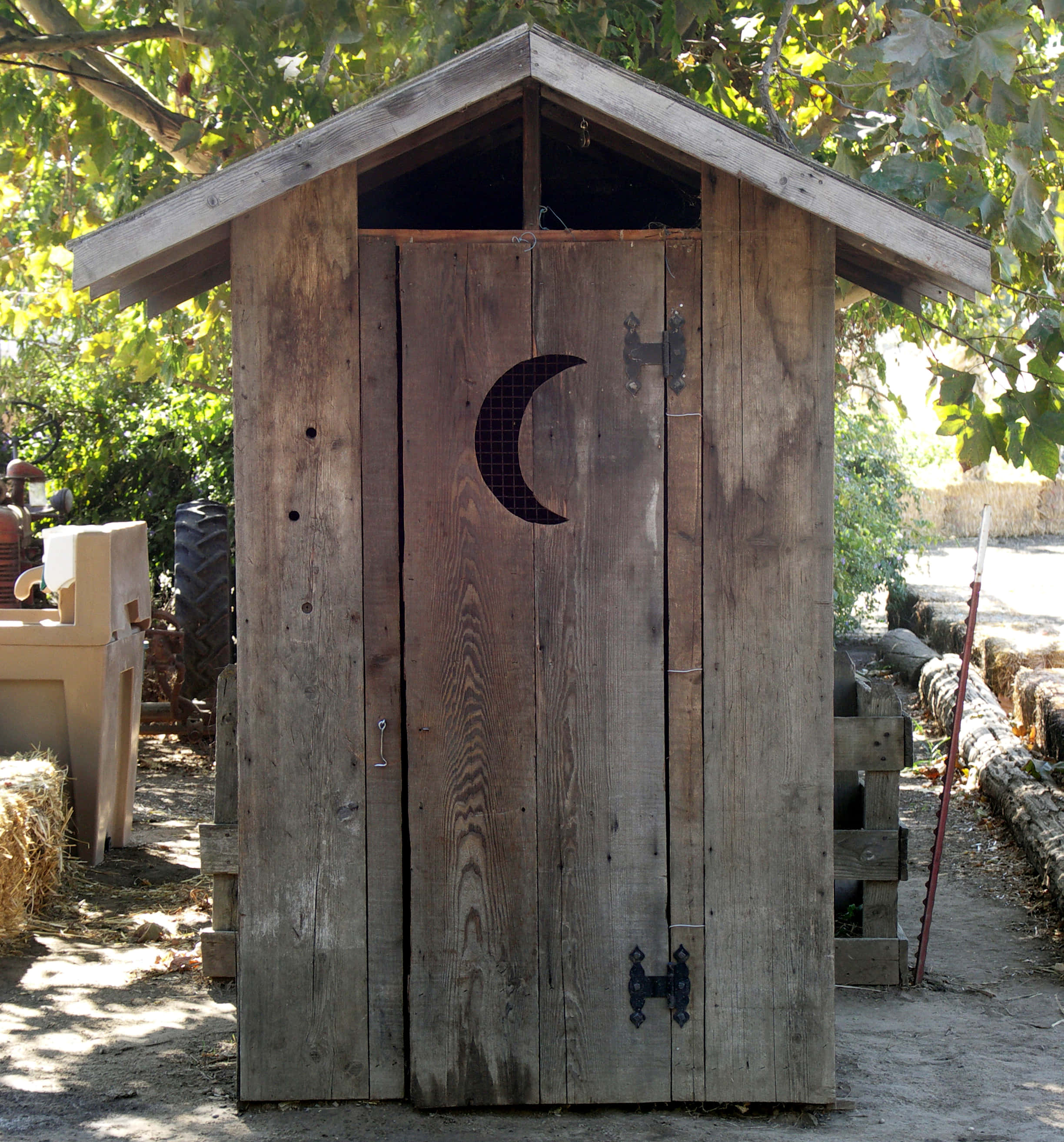 Outhouse 桌布 壁紙 — 一座質樸的木製戶外廁所，帶有新月形鏤空門，矗立在陽光明媚的鄉村環境中枝葉繁茂的樹木下。